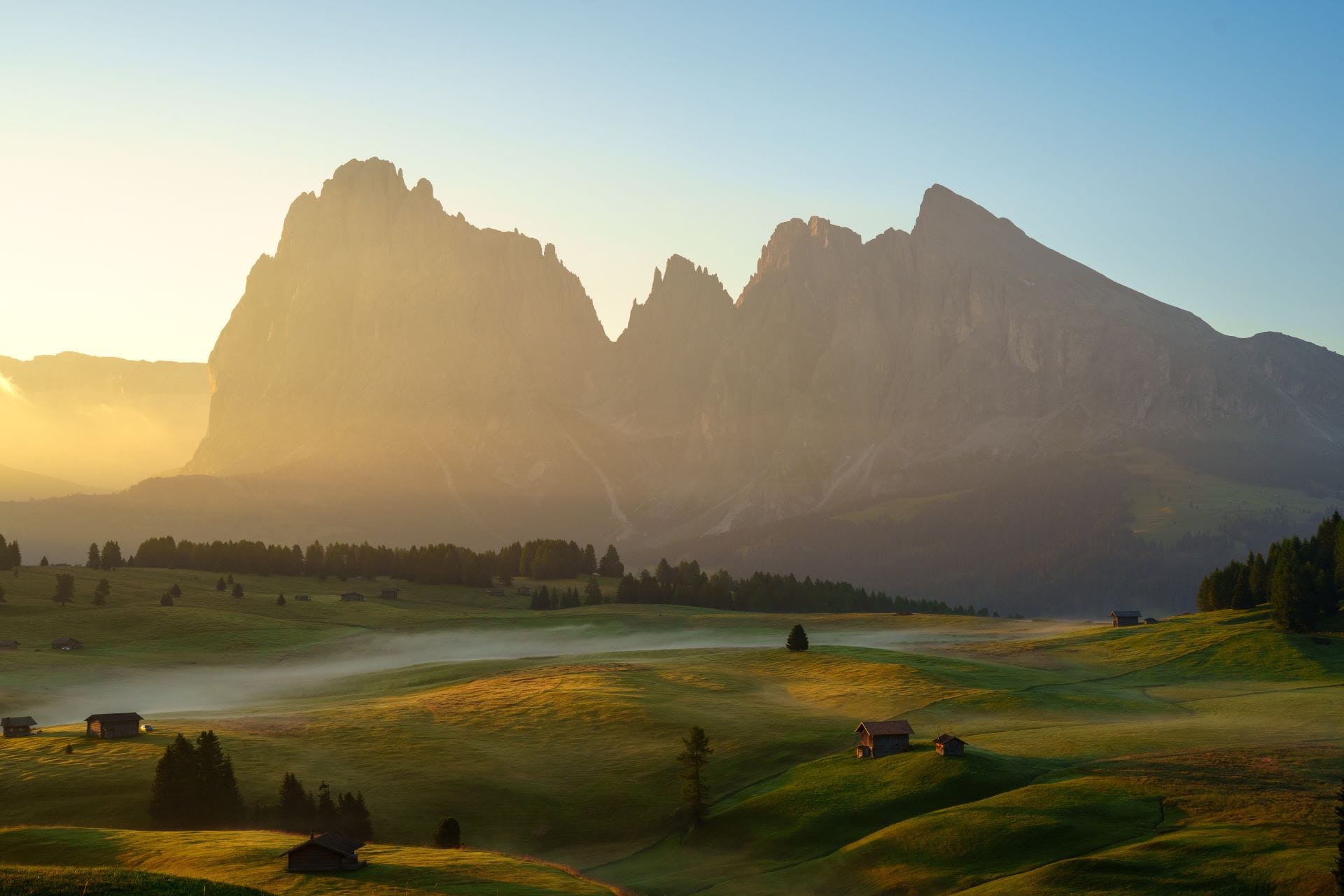 Il massiccio del Sassolungo illuminato dalla luce dorata dell'alba, con una leggera nebbia mattutina che accarezza i prati verdi e le tradizionali baite in legno dell'Alpe di Siusi