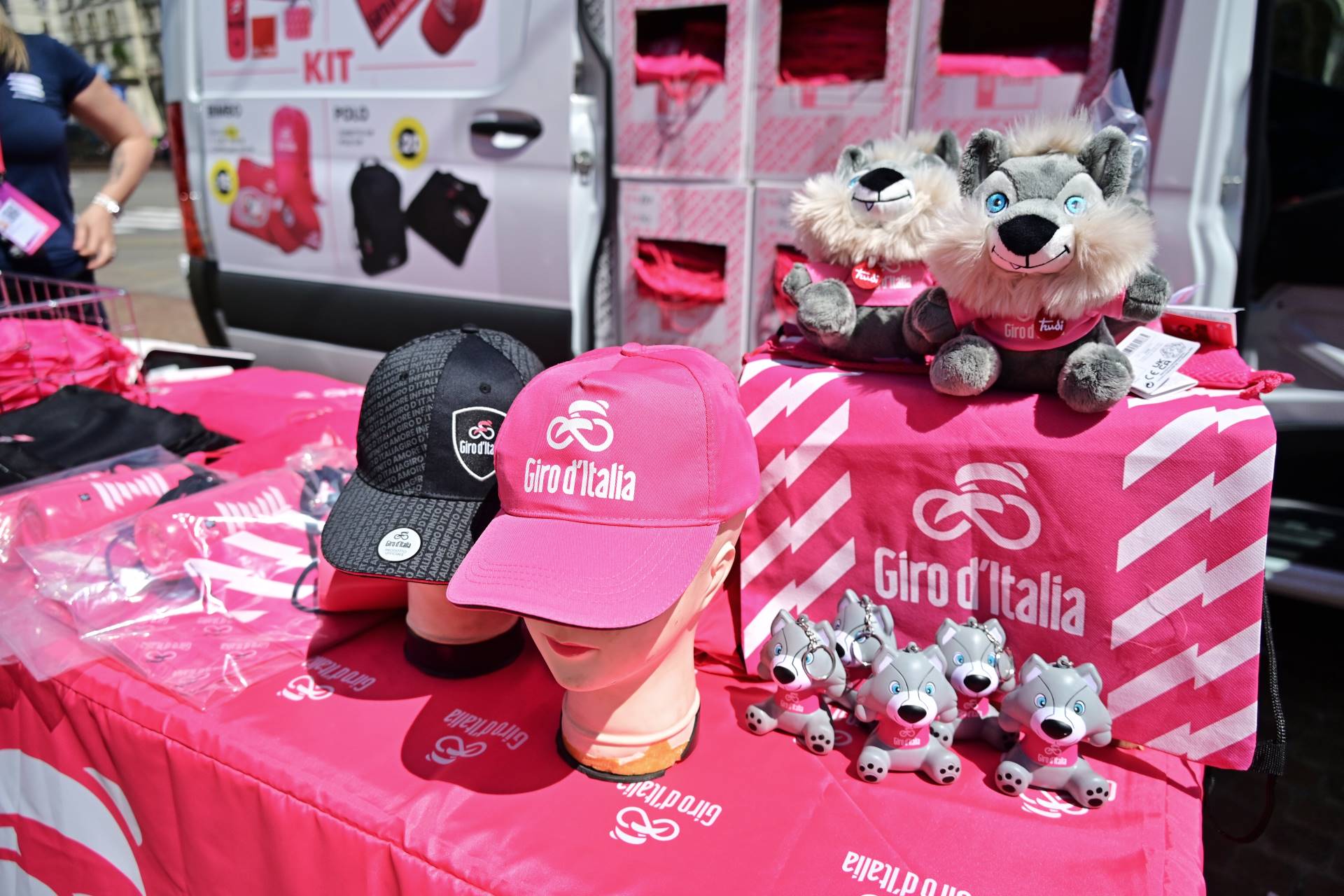 Vendor stand displaying official Giro d'Italia merchandise including pink hats, mascot plush toys, and cycling souvenirs.