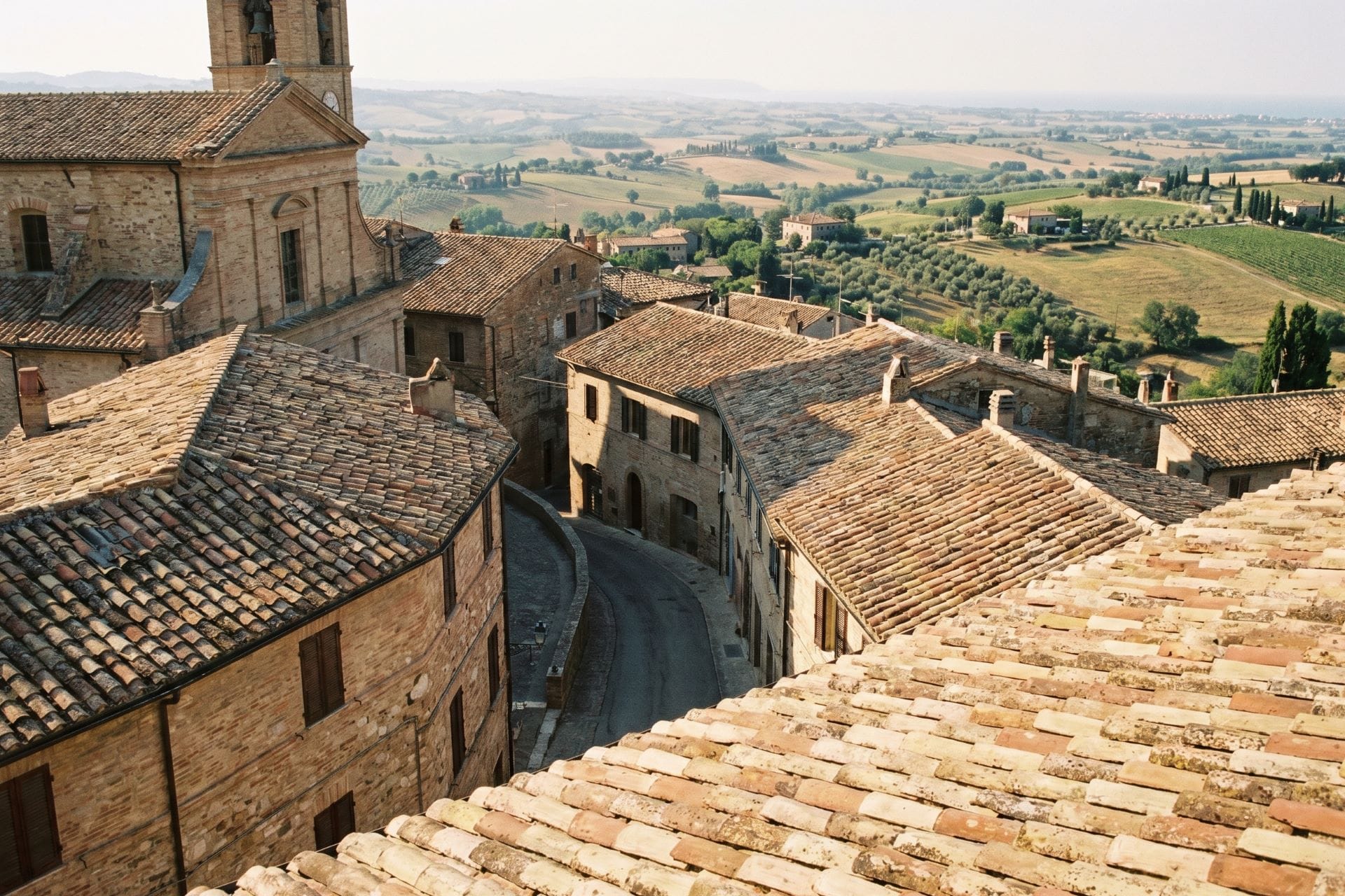 Elevated view over terracotta rooftops of a medieval hill town borgo with rolling agricultural landscape in the background, central Italy, summer afternoon