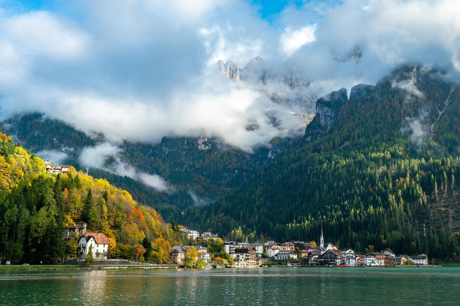 Panoramic summer view of Lago di Alleghe reflecting the surrounding towering peaks of the Italian Dolomites under a clear blue sky.