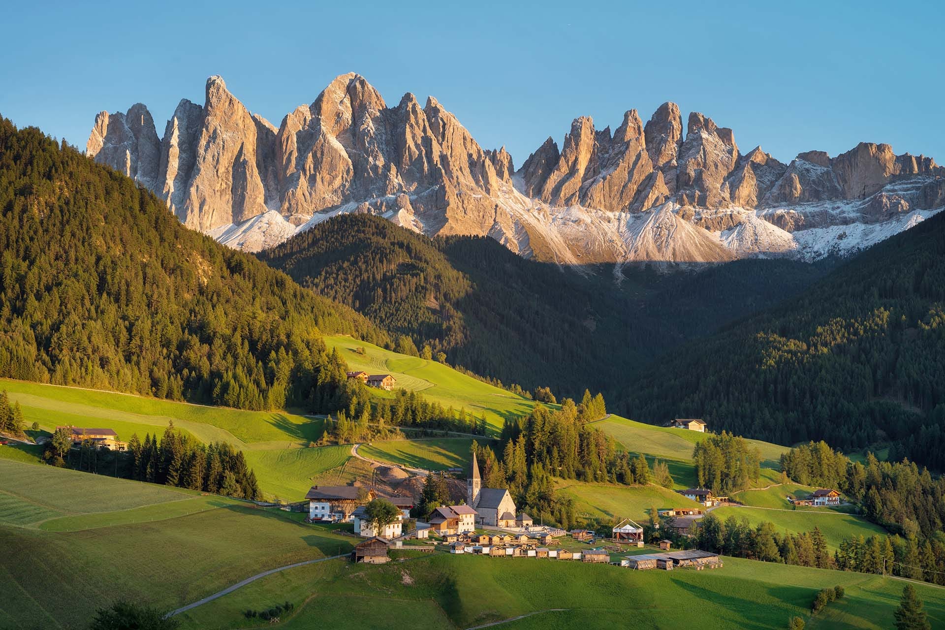 A traditional Alpine village with wooden chalets nestled in a green valley at the foot of the dramatic Odle (Geisler) peaks in the Dolomites, South Tyrol, Italy, on a clear summer day.