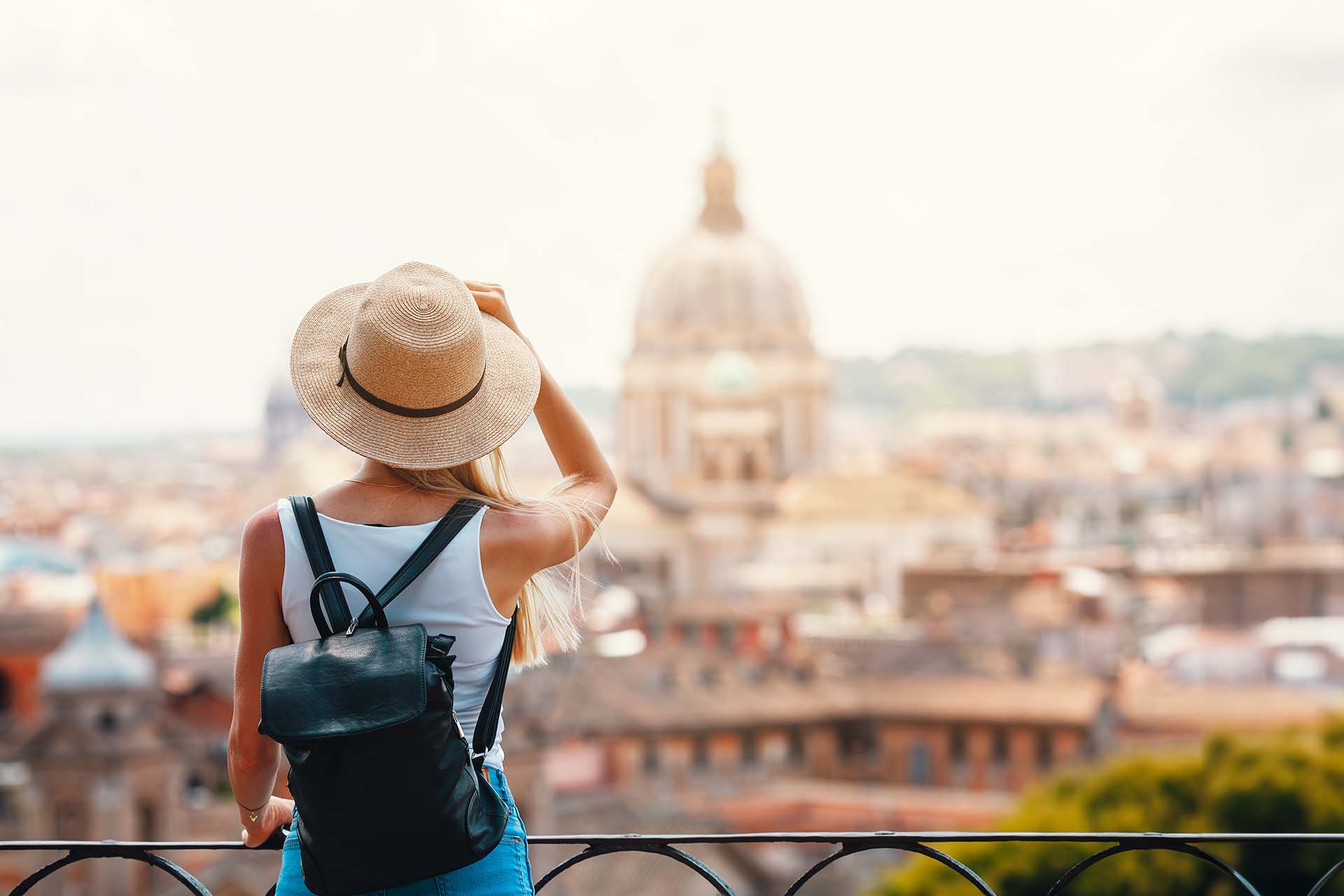 A young female traveler with a straw hat and backpack seen from behind, gazing over the rooftops and domes of Rome on a summer day, Italy.