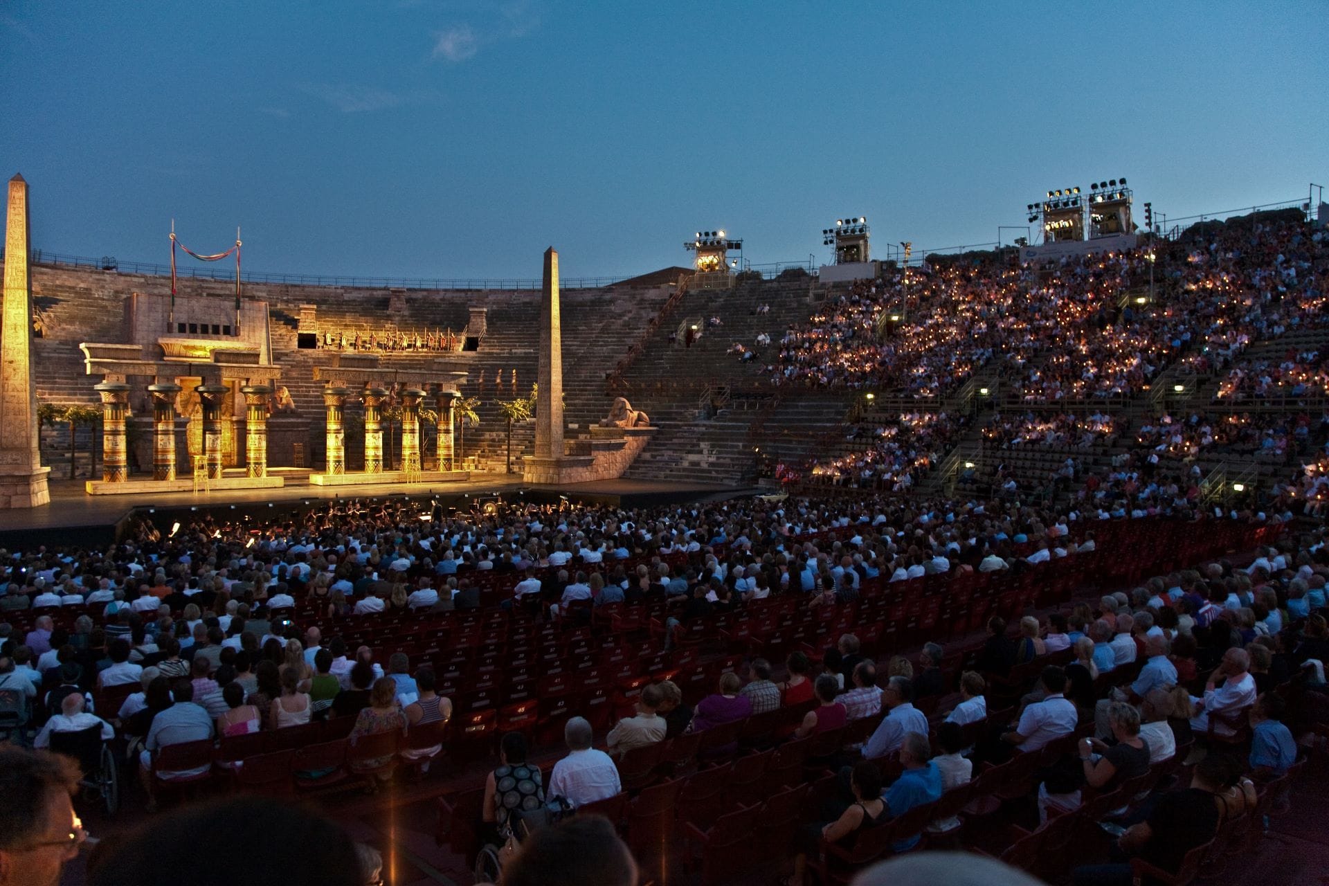 Spectators lighting candles in the tiered stone seats of the Arena di Verona at dusk before an opera