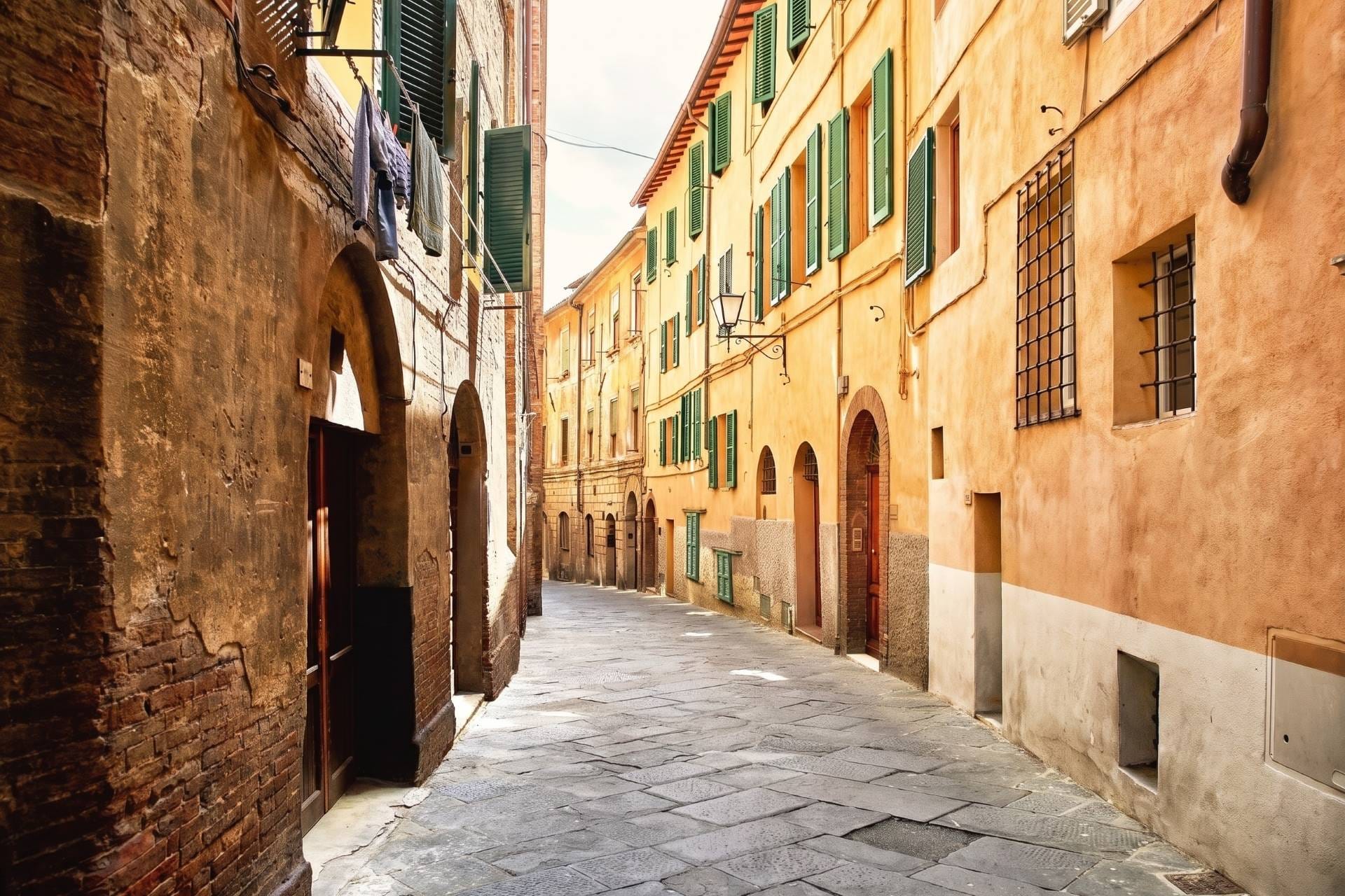 Sunlit ochre Siena backstreet with green shutters and hanging laundry in the walled historic center