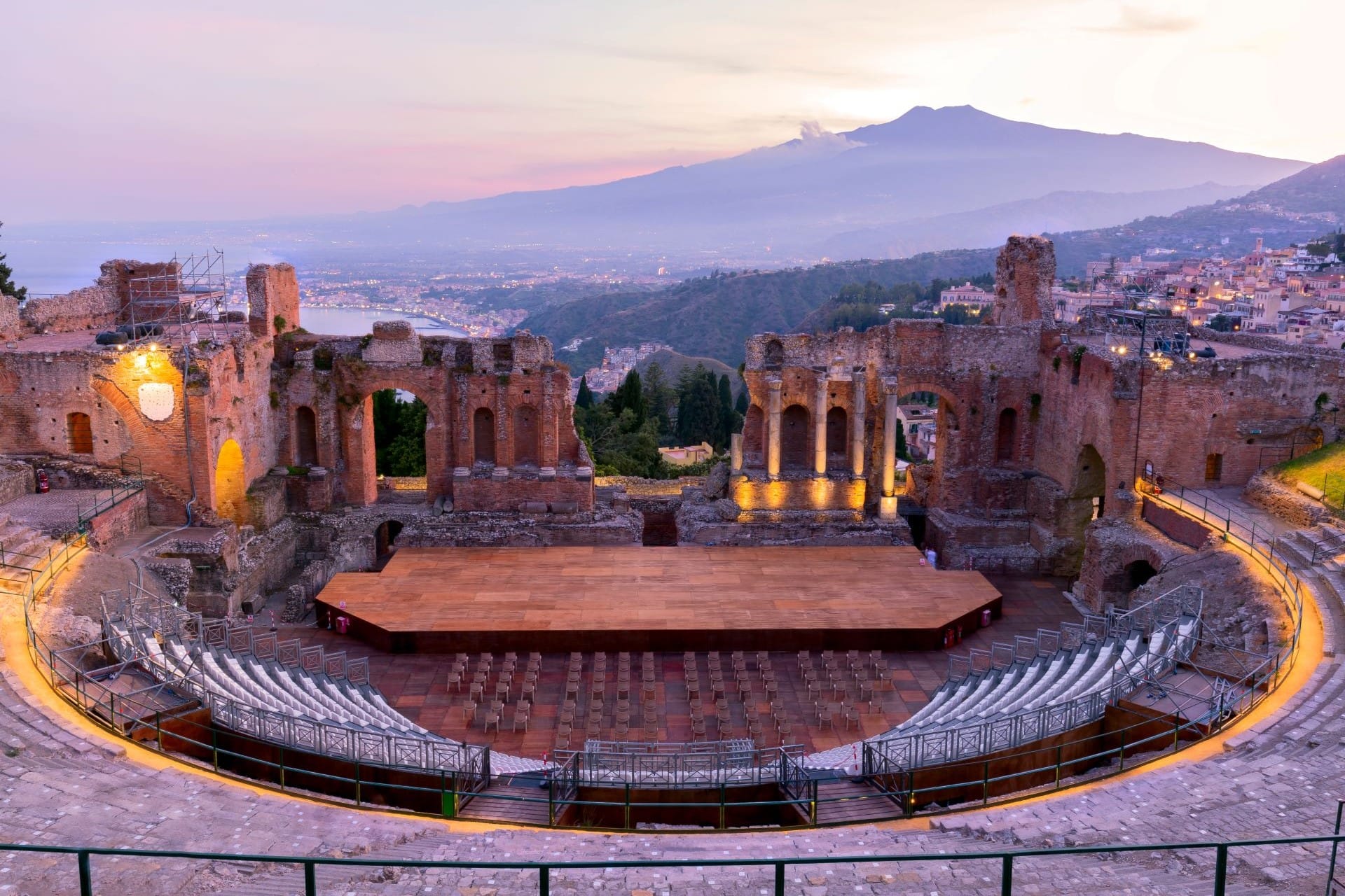 The ancient Greek theater of Taormina framing Mount Etna and the bay of Giardini Naxos at dusk in Sicily