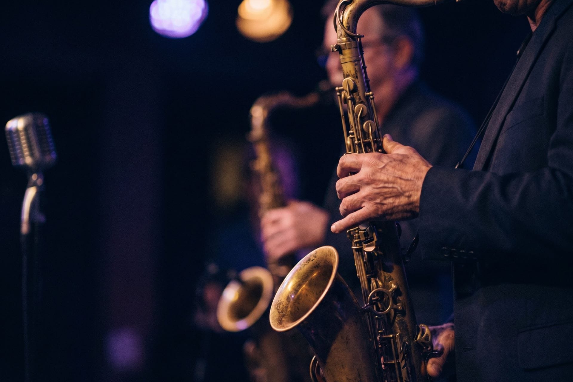 A musician's hands on a tenor saxophone under blue stage light beside a vintage microphone at a jazz set