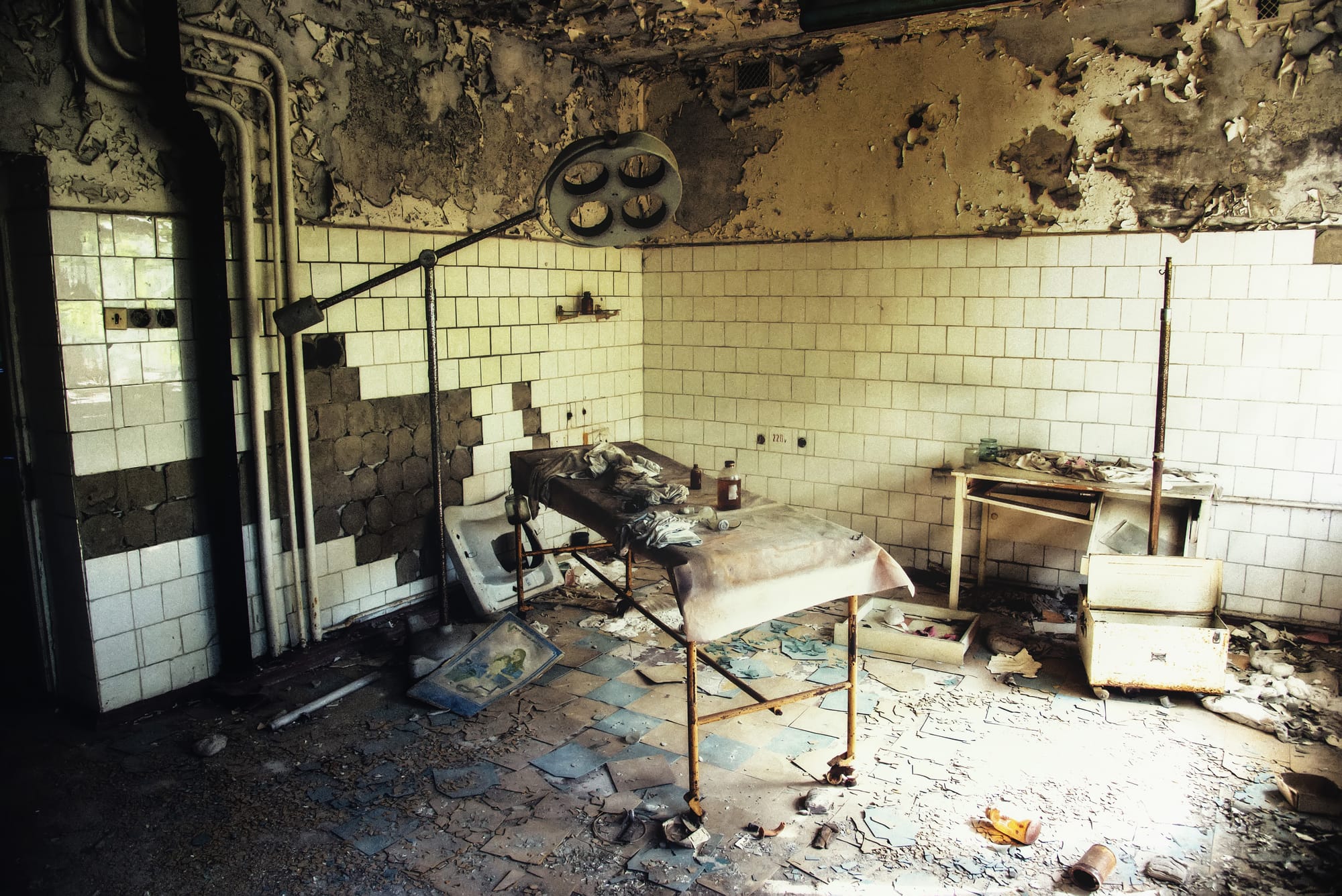 Abandoned examination room in Pripyat Hospital, showing severe decay with peeling walls, broken white tiles, and a rusty medical table with overhead surgical light. Debris covers the floor of this Chernobyl disaster relic.