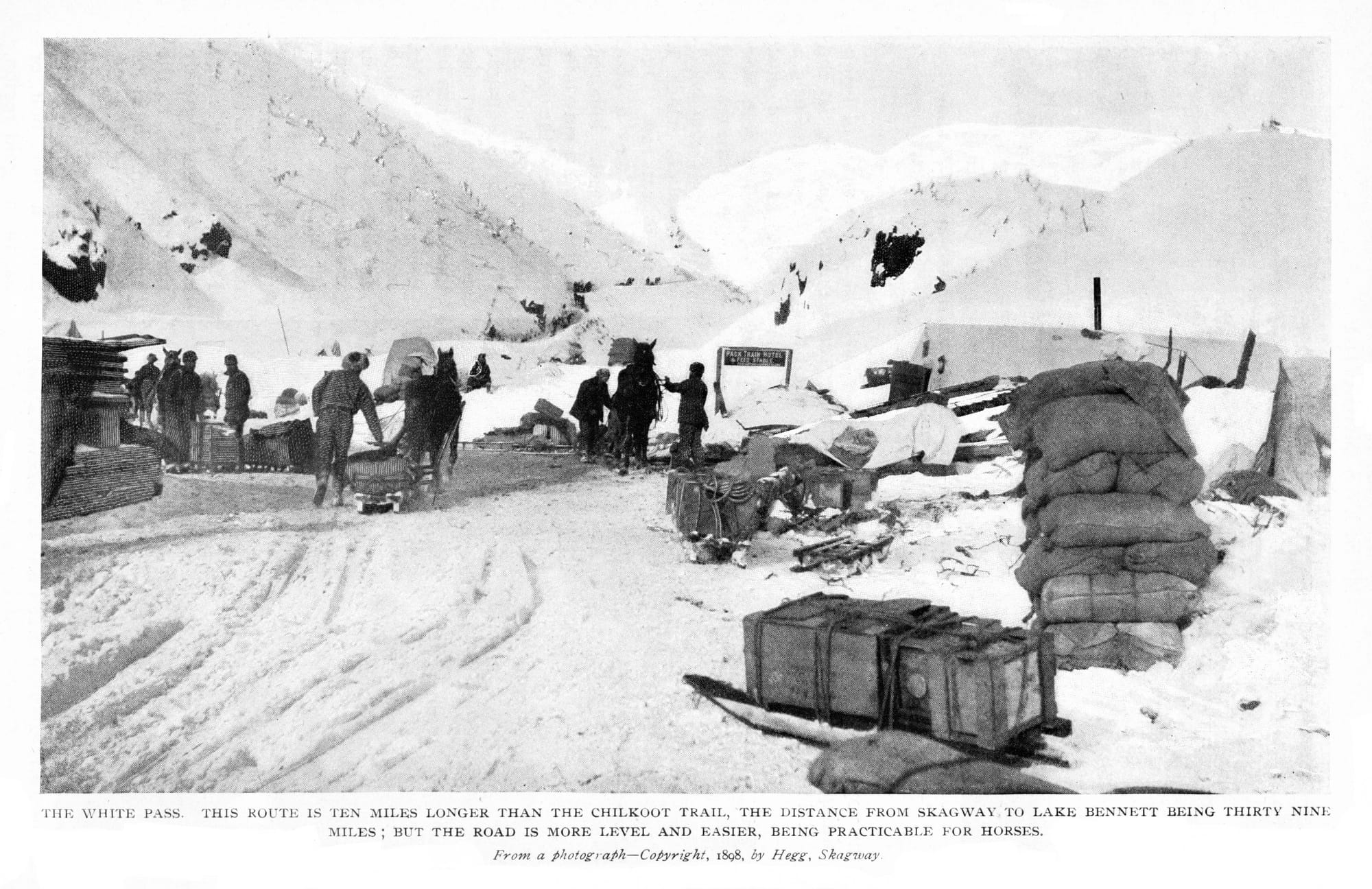 Historic black and white photo from 1898 showing White Pass during the Klondike Gold Rush. People and horses navigate through snowy terrain with supplies and makeshift structures amid mountainous landscape.