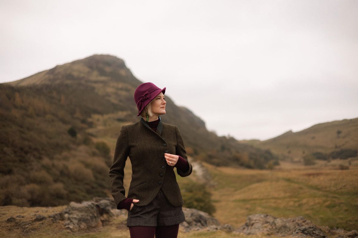 Caitlin wearing a purple felt hat, green Walker Slater jacket, standing in front to Arthur's Seat.
