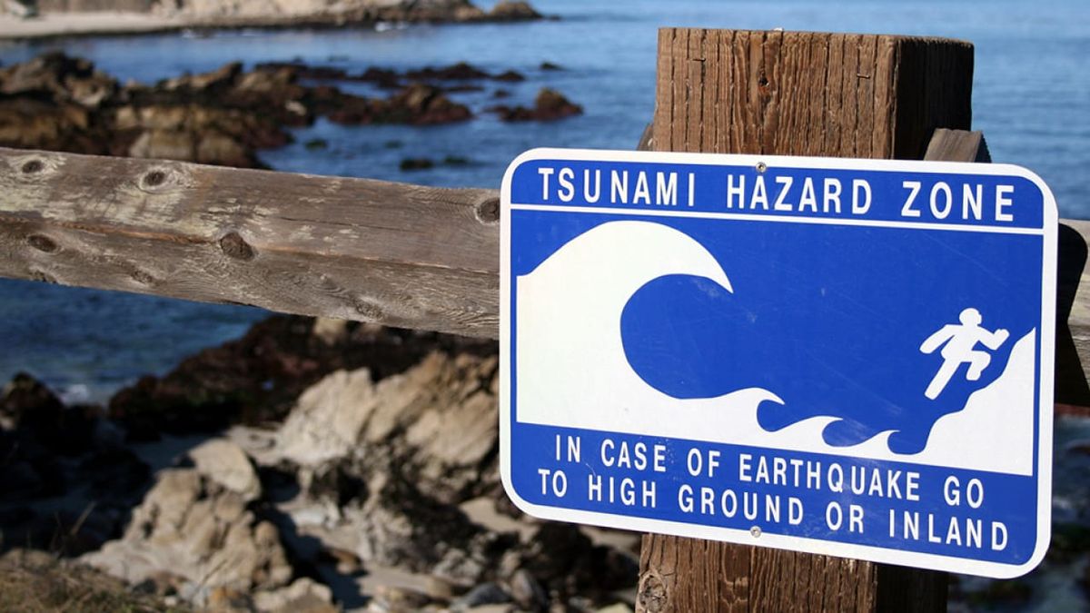 Blue and white Tsunami Hazard Zone sign on a fence by the ocean, directing people "in case of earthquake go to high ground or inland."