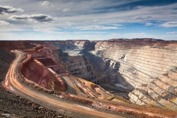 Massive open-cut gold mine with terraced layers carved into earth. Winding dirt road spirals down left side. Tiny equipment at bottom shows enormous scale.