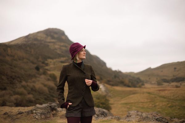 Caitlin wearing a purple felt hat, green Walker Slater jacket, standing in front to Arthur's Seat.