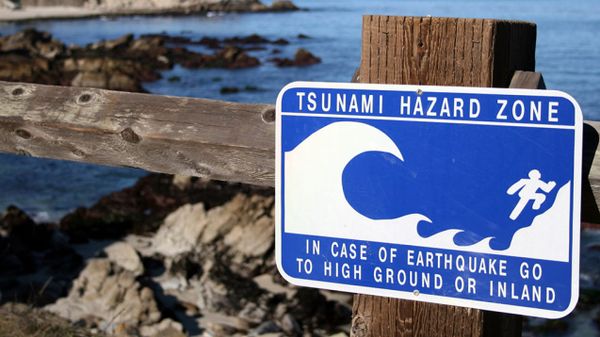 Blue and white Tsunami Hazard Zone sign on a fence by the ocean, directing people "in case of earthquake go to high ground or inland."