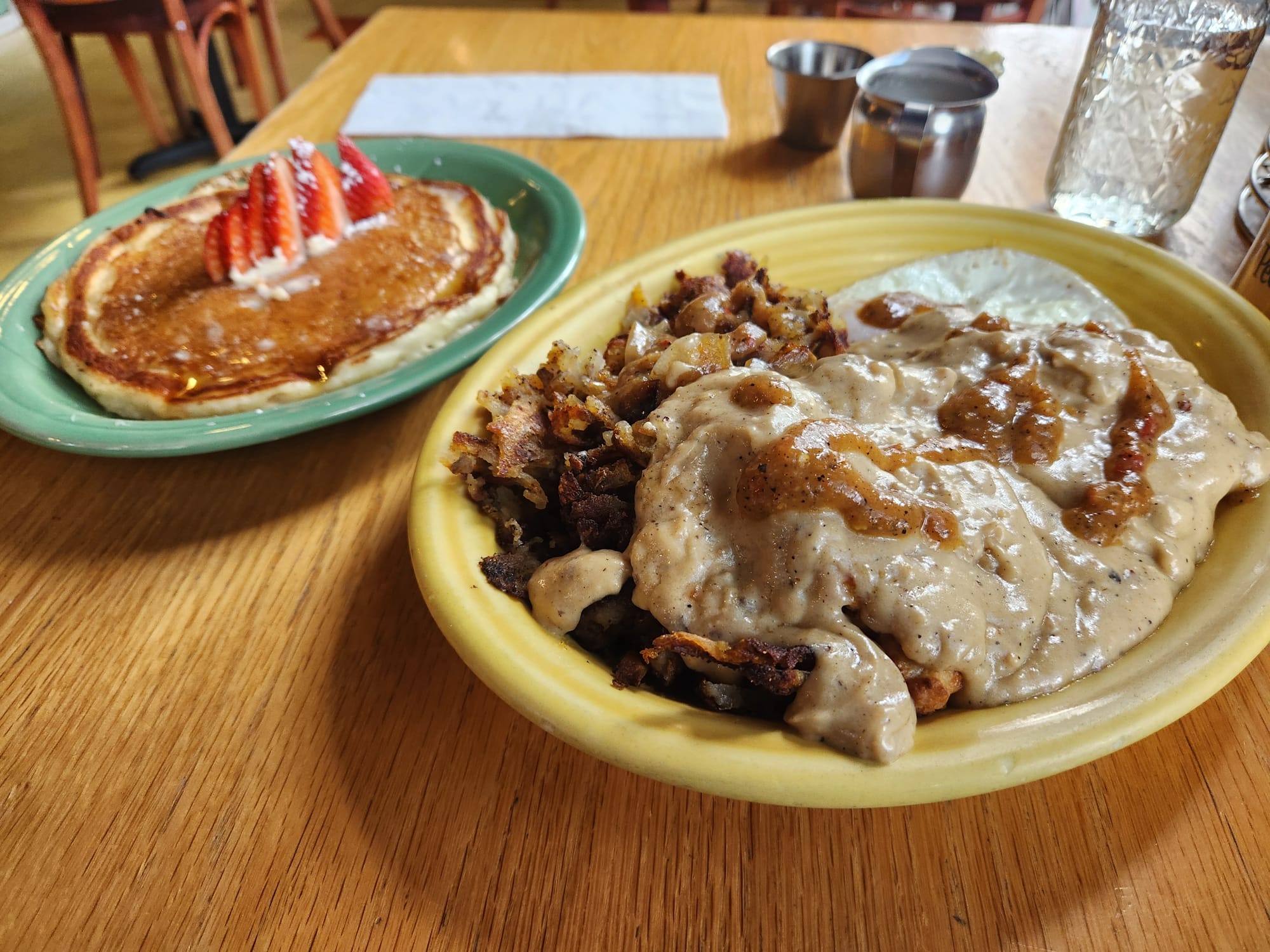 A plate of biscuits and gravy with eggs over easy at Little Cheerful Cafe, with one hotcake on a sideplate with strawberry garnish..