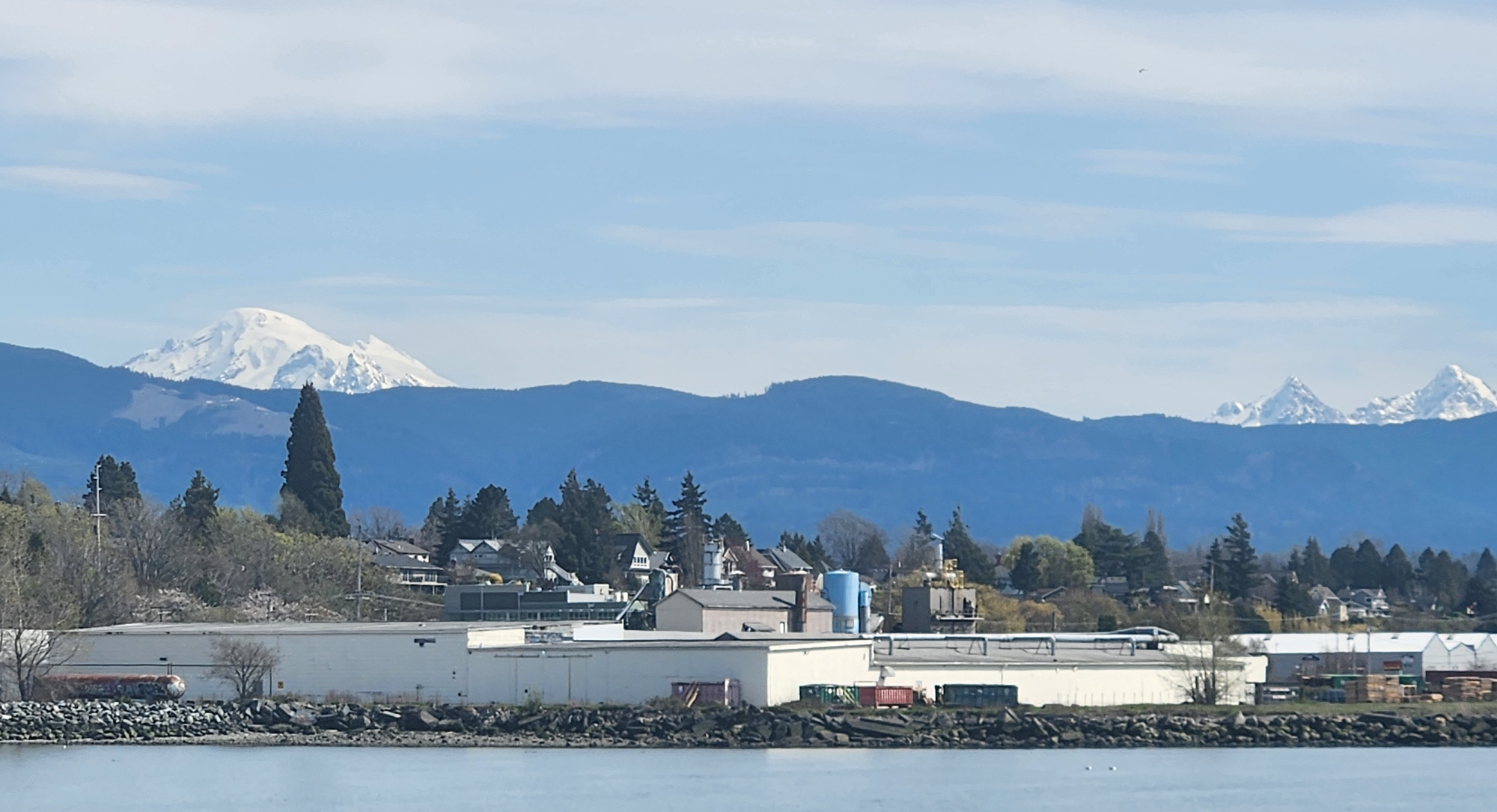 The snowy peak of Koma Kulshan peeks above the foothills beyond Bellingham, with a waterside warehouse in the foreground..