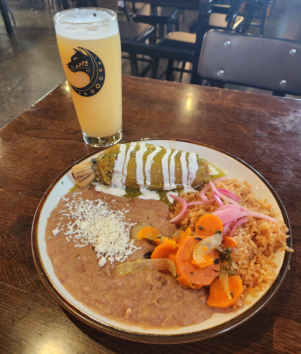 A tamale covered in green salsa and crema on a plate with rice, beans and pickled onions and carrots.