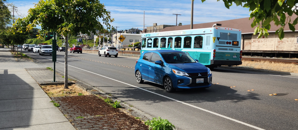 A light-blue colored WTA bus travels west on a street with a small car in the adjacent lane traveling east.