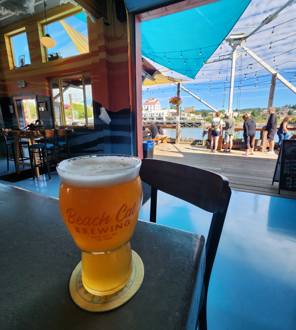A glass of beer on a bar with an open garage door opening onto a waterside deck with five people standing near a railing.
