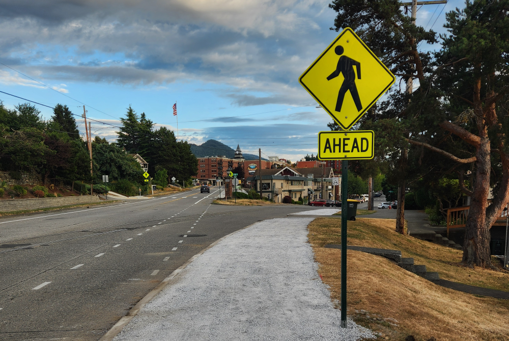 A compacted gravel pathway along the west side of of a roadway with a yellowish pedestrian crossing "ahead" sign.gn