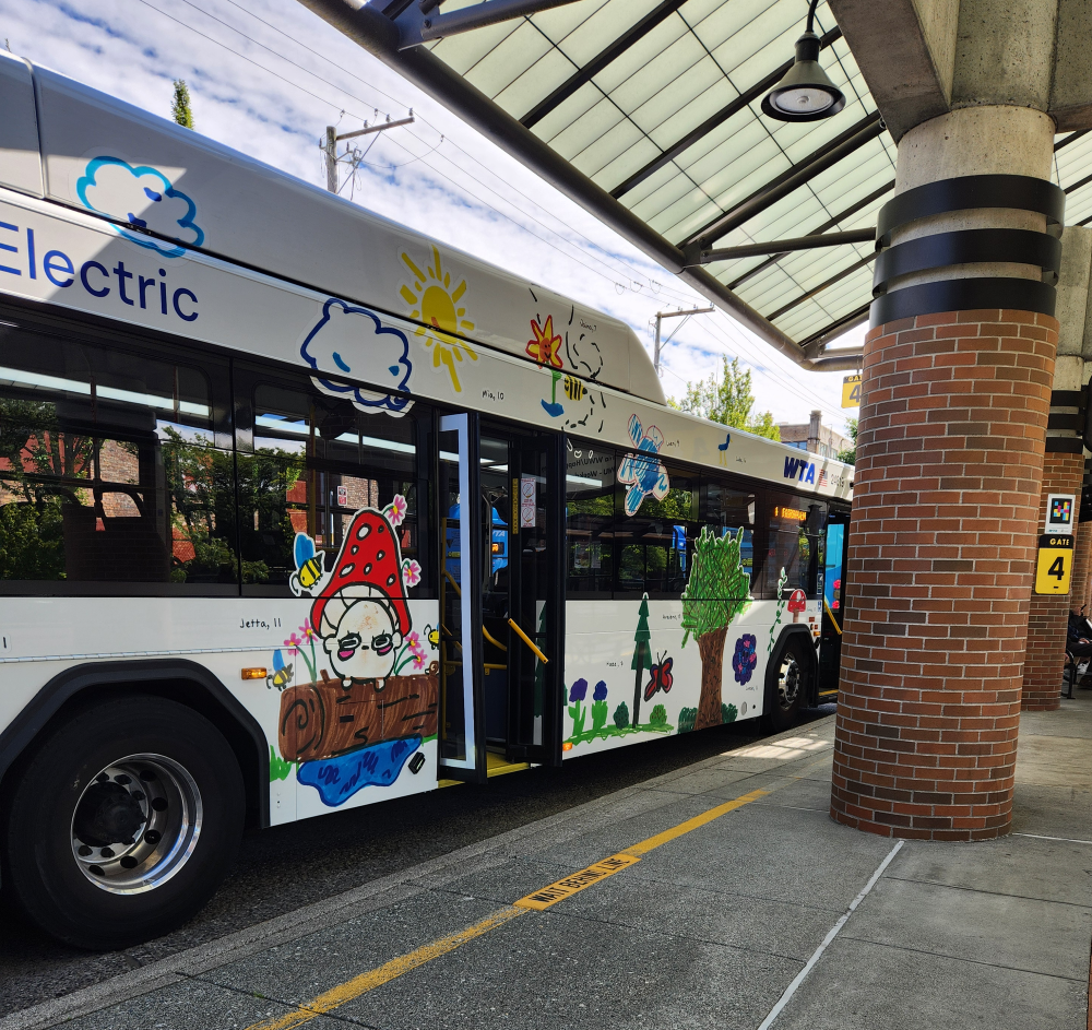 A bus with a exterior wrap featuring children's art sits at a bus bay in a transit center.