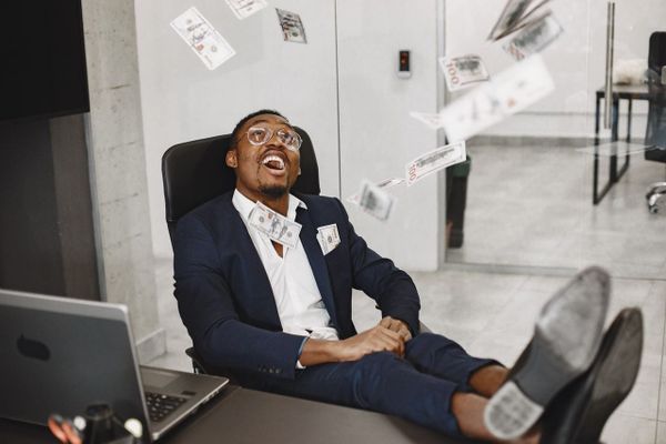 Man sat in chair laughing at falling money with his feet on a desk. 