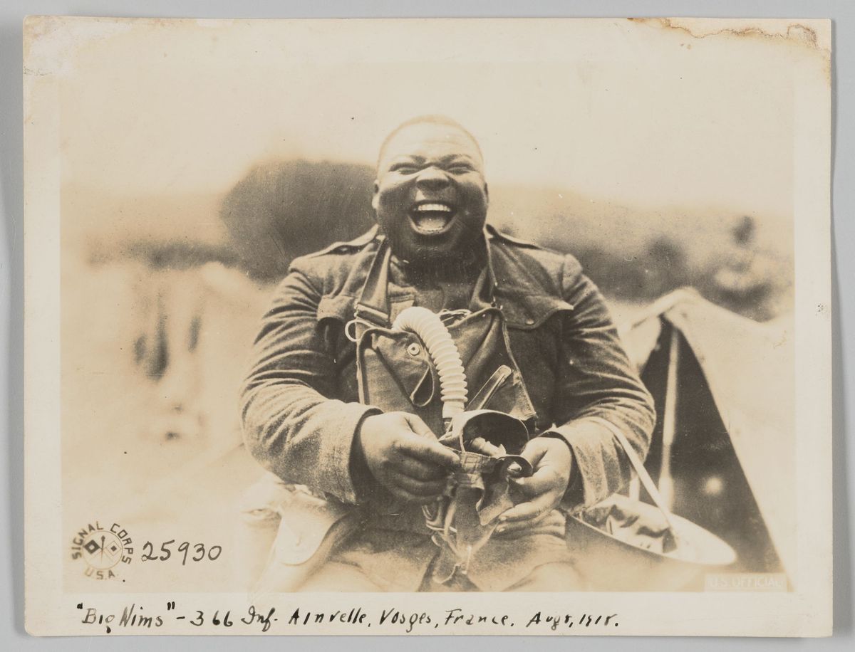 A sepia-toned picture of a man laughing with a gas mask in his hands. The photo is dated “Ainvelle, France, Aug 8, 1918.”