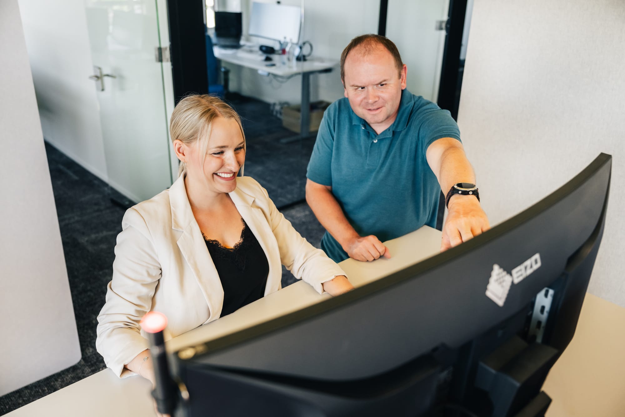 SEO-Spezialistin und Analytics-Spezialist besprechend stehnend am Arbeitsplatz eine Analyse. Er zeigt auf den Bildschirm.