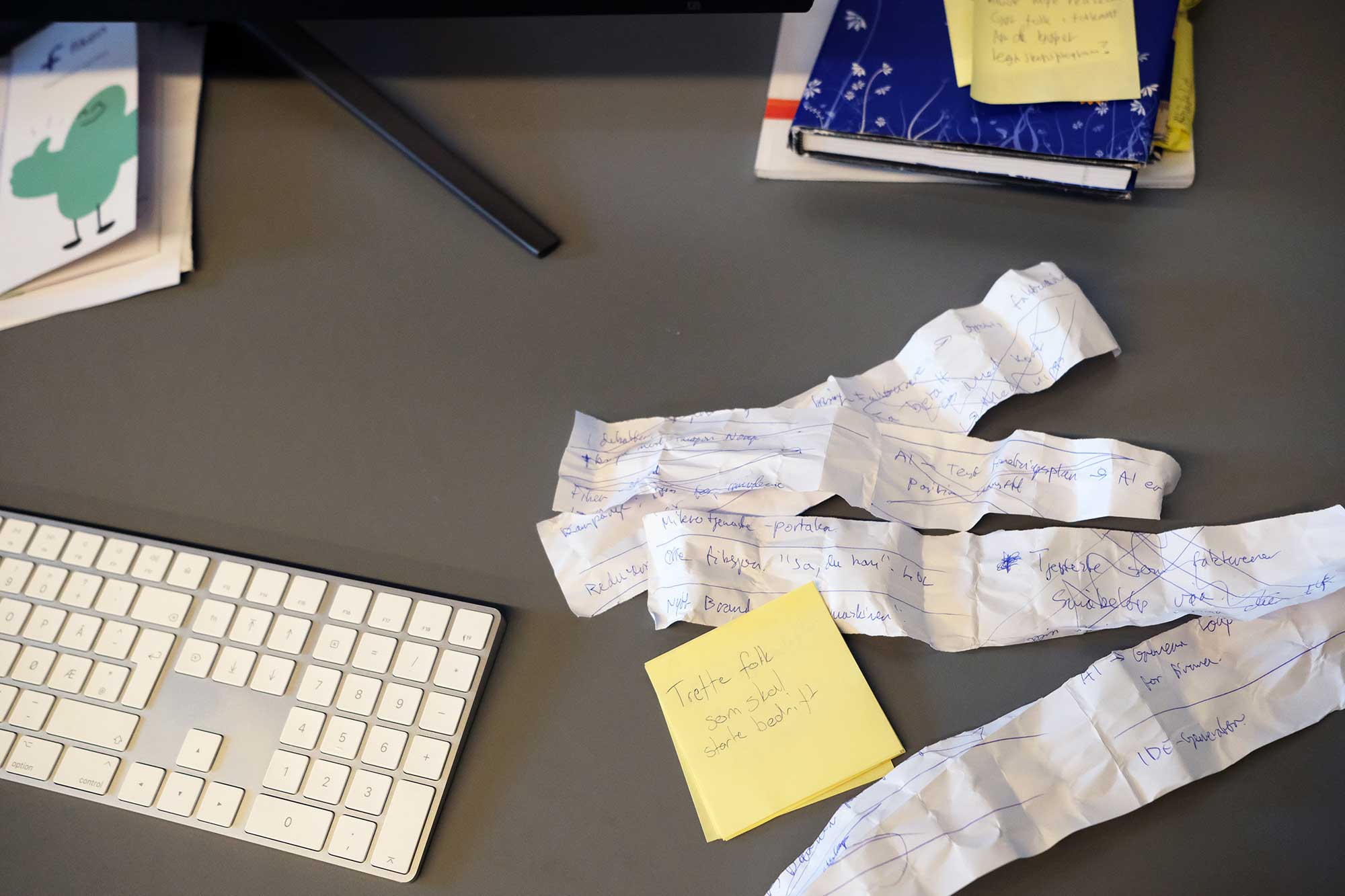 Foto av et skrivebord med tastatur, bøker og post it-lapp på.