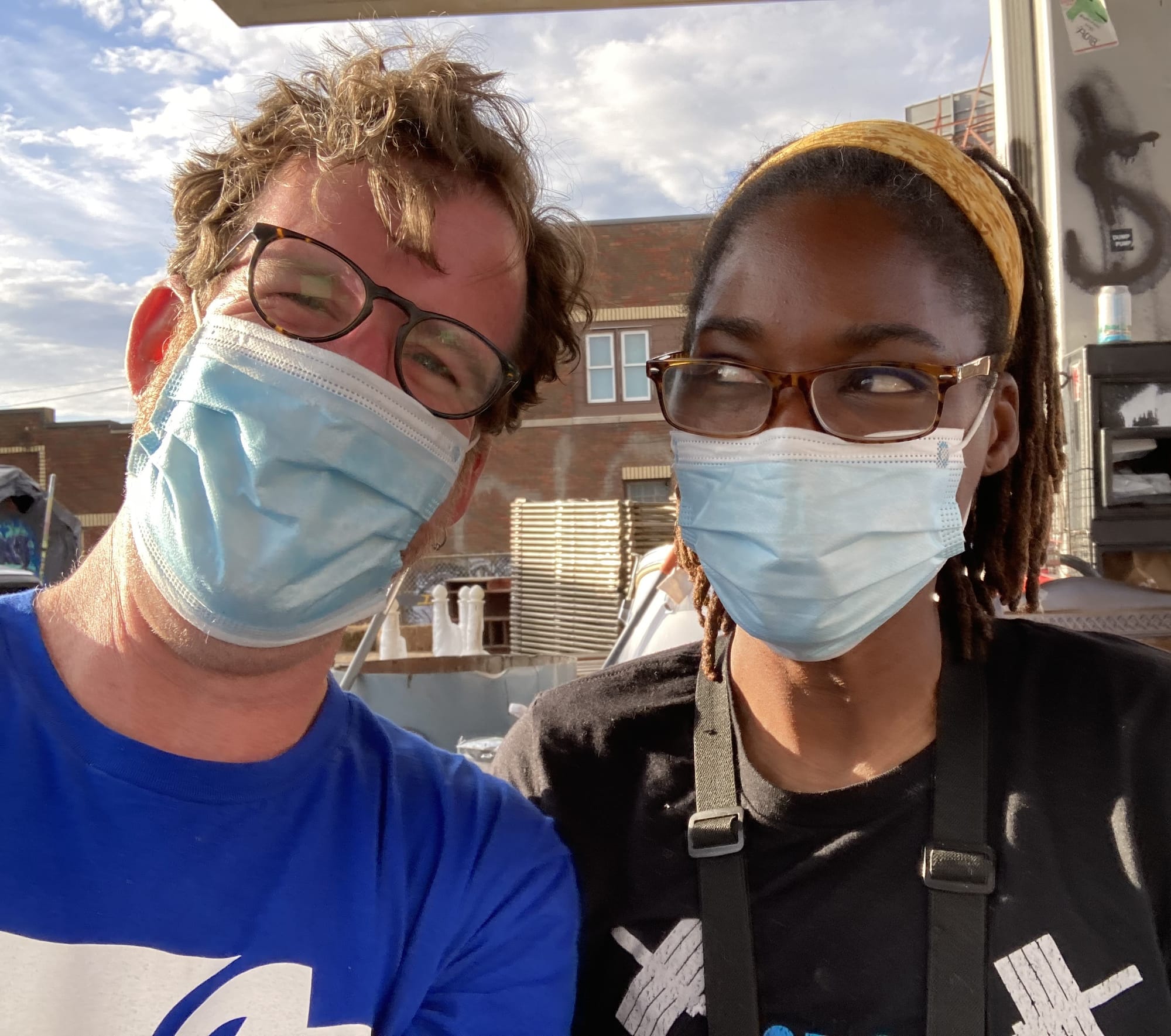 A white man in blue shirt looks into the camera while an African-American woman looks playfully at the man. Both are wearing medical masks.