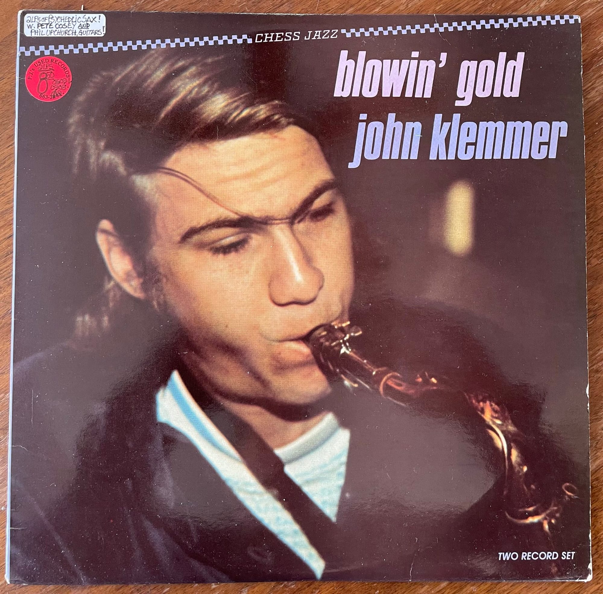 The album cover for John Klemmer's "Blowin' Gold", featuring a photo of the musician, a young white man with blond hair in a white shirt and black sweater blowing a saxophone.