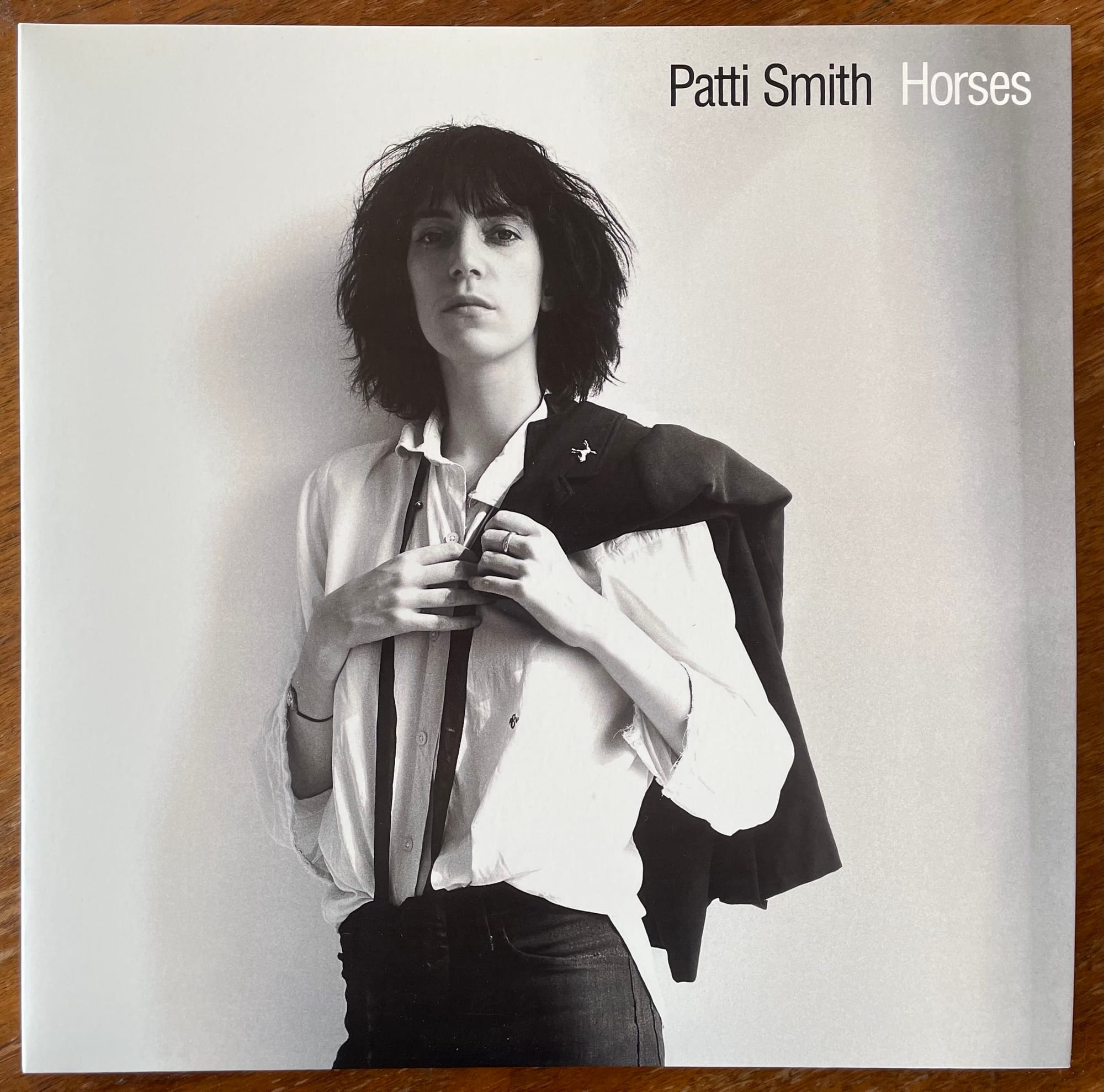 Album cover of Patti Smith's Horses album, a black and white photo of Patti looking at the camera, an androgynous look of white shirt, black slacks, suspenders, jacket slung over the shoulder. Her shoulder-length bob is messy.