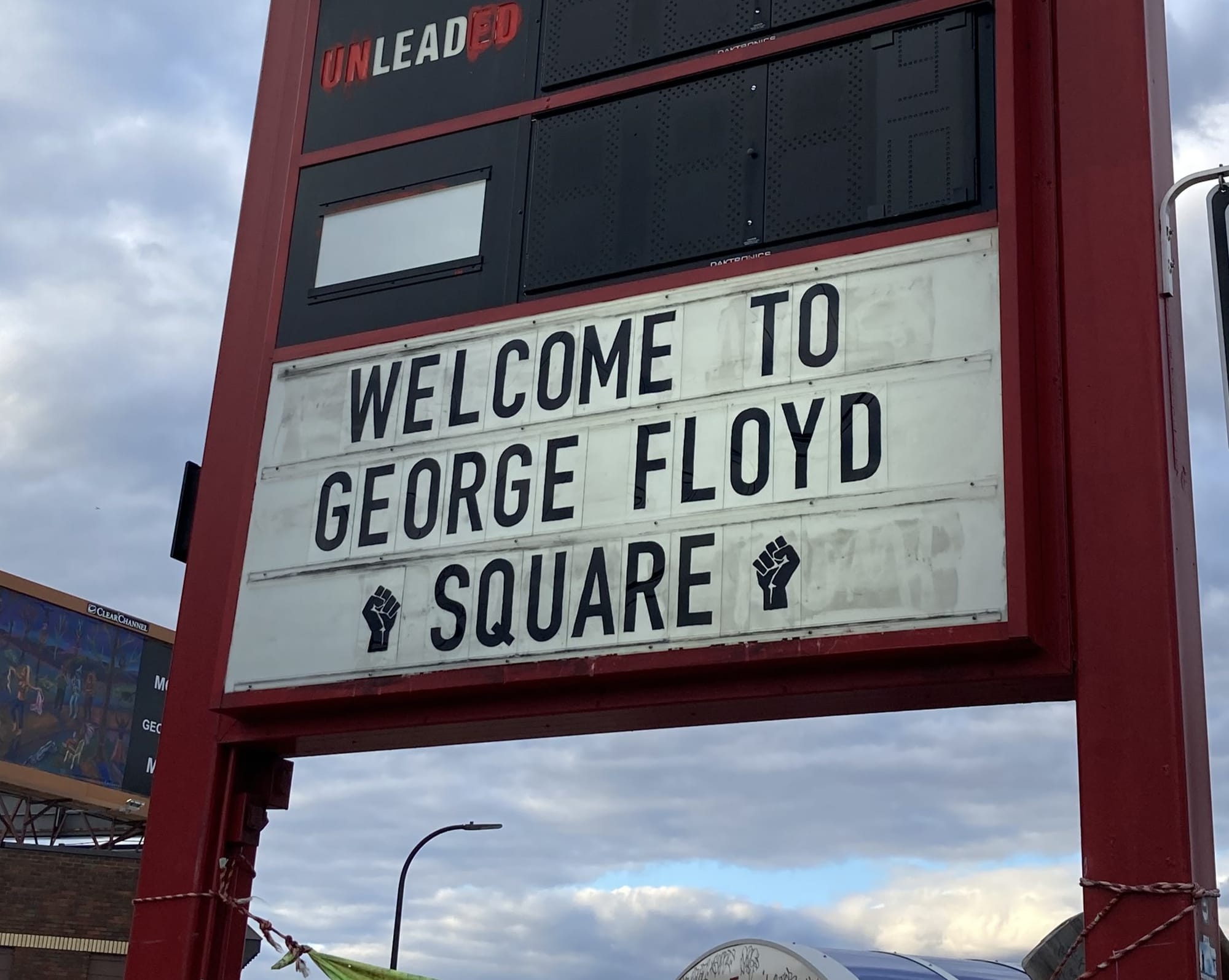 Against a cloudy sky, a gas station marquee reads "Welcome to George Floyd Square." Black power fists are on other side of the word "Square."