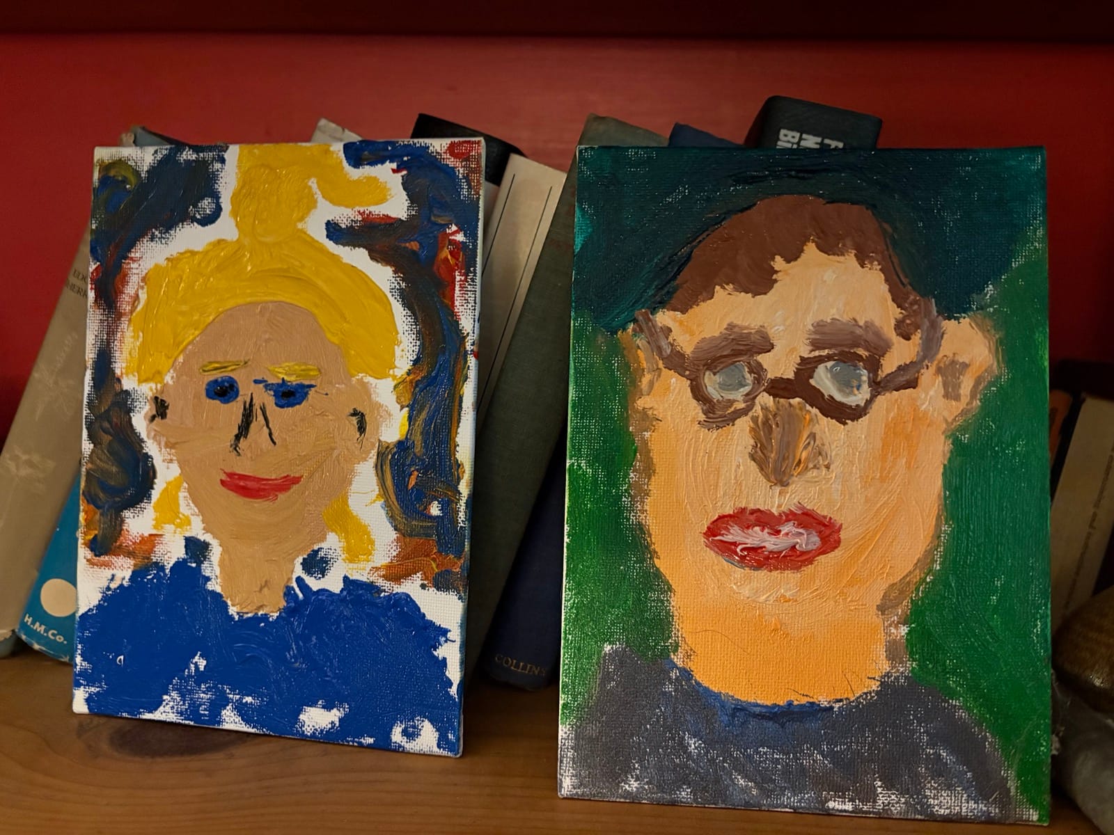 Two brightly colored portraits resembling people, possibly painted by raccoons?, sit on a bookshelf.