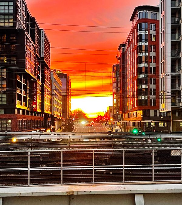 A sunrise of brilliant gold, orange, and pink appears over a street and train tracks between tall buildings.