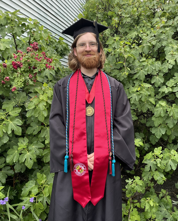 A handsome man with beard and long hair wears an academic robe, mortarboard and tassle, a red stole, and braided cords in front of a fig tree.