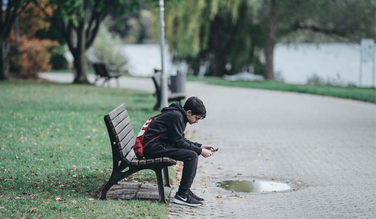 A teenager on a park bench scrolls on his phone.