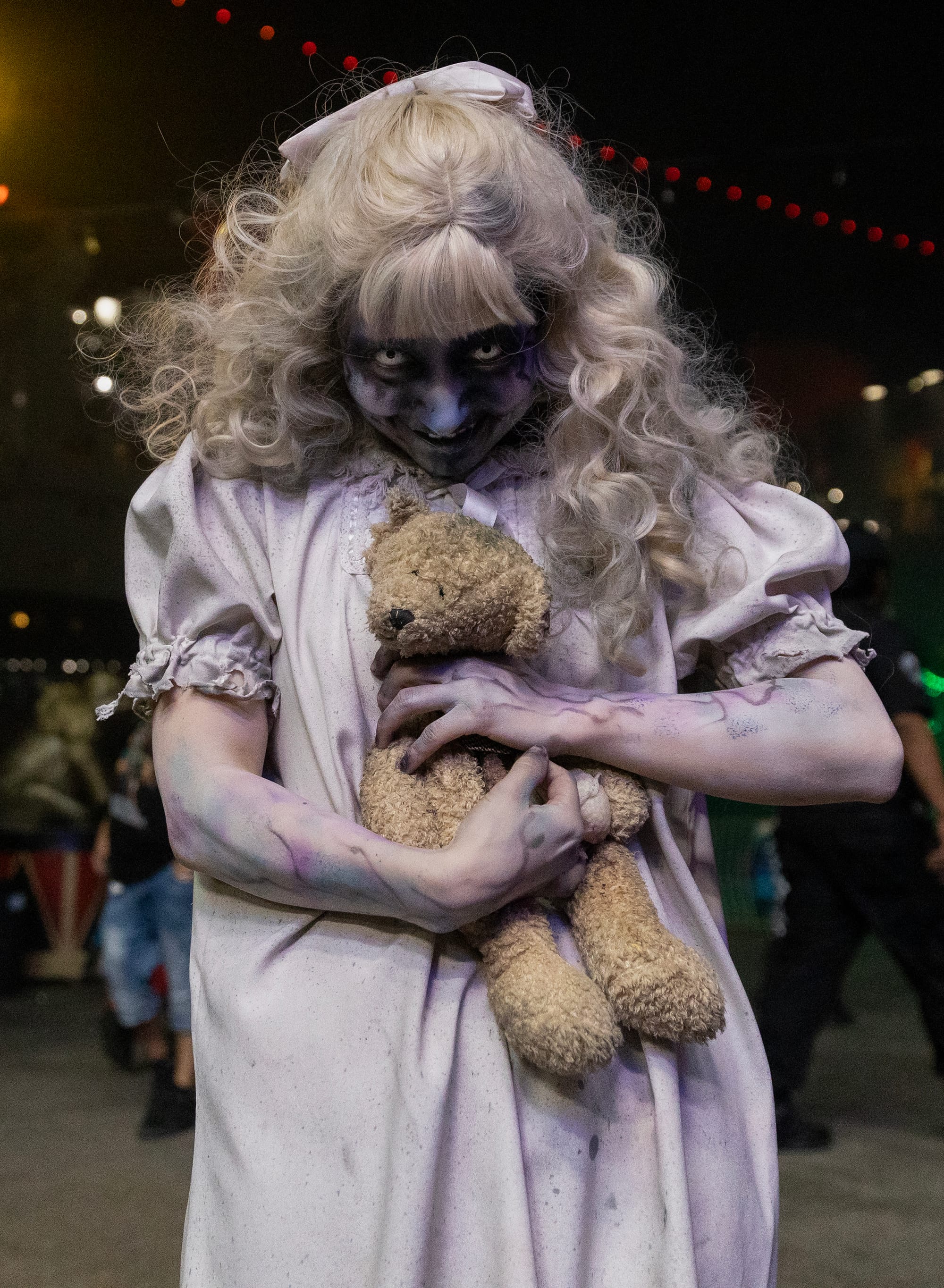 A woman in heavy horror makeup and a white dress clutches a teddy bear.