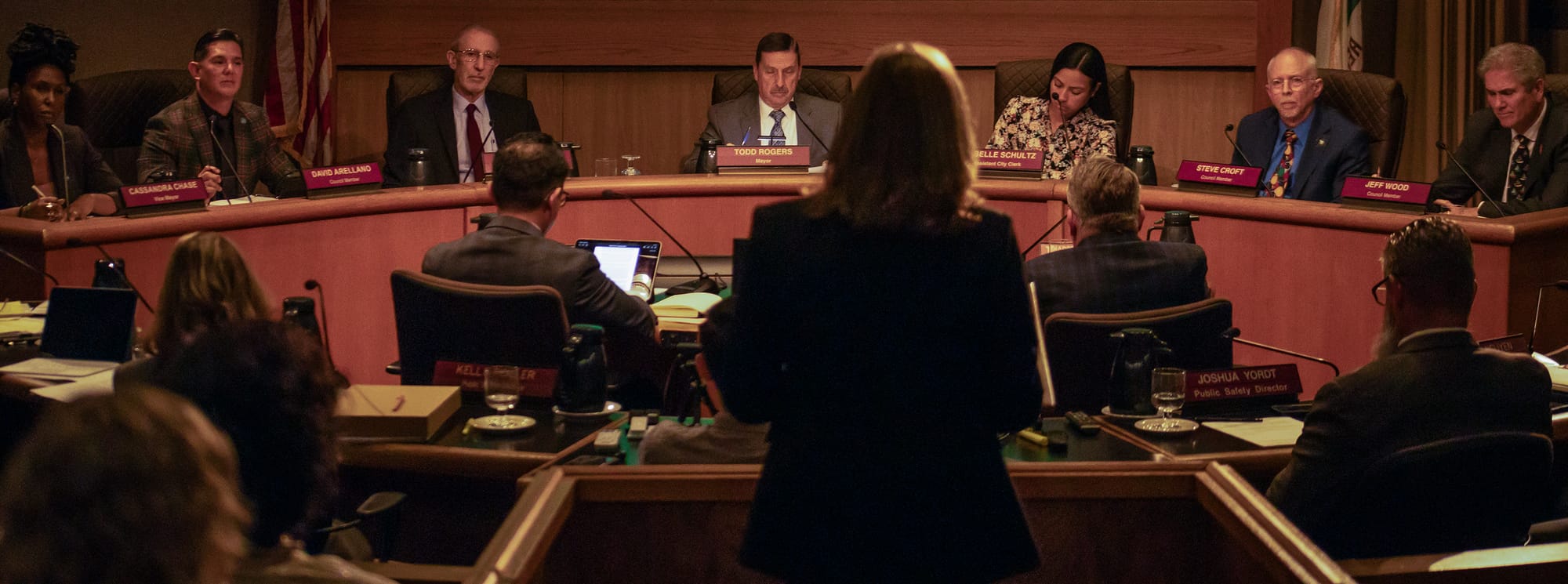 The shadowy outline of a woman stands before seven people seated at a dais.