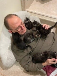 Five kittens walk on a man's chest and shoulders.