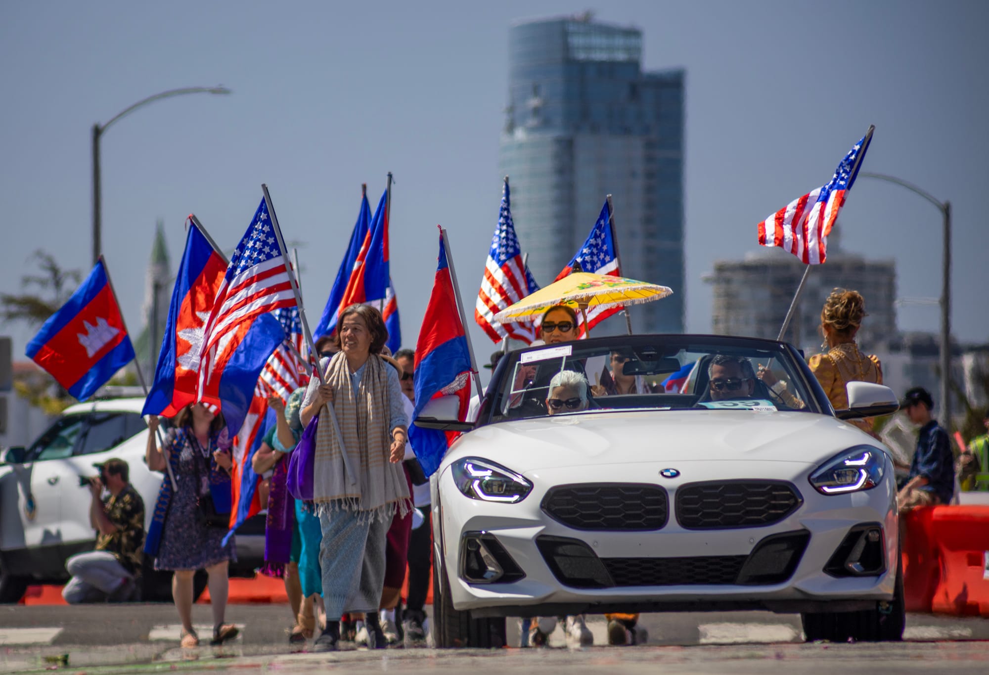 People hold flags while walking next to a white car.
