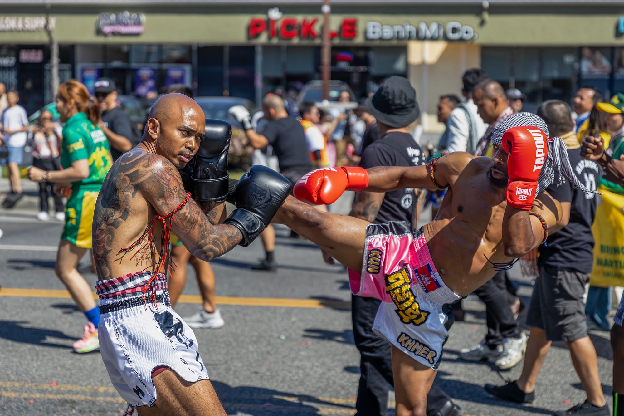 Two men in white shorts box each other in the middle of the street as people walk by.