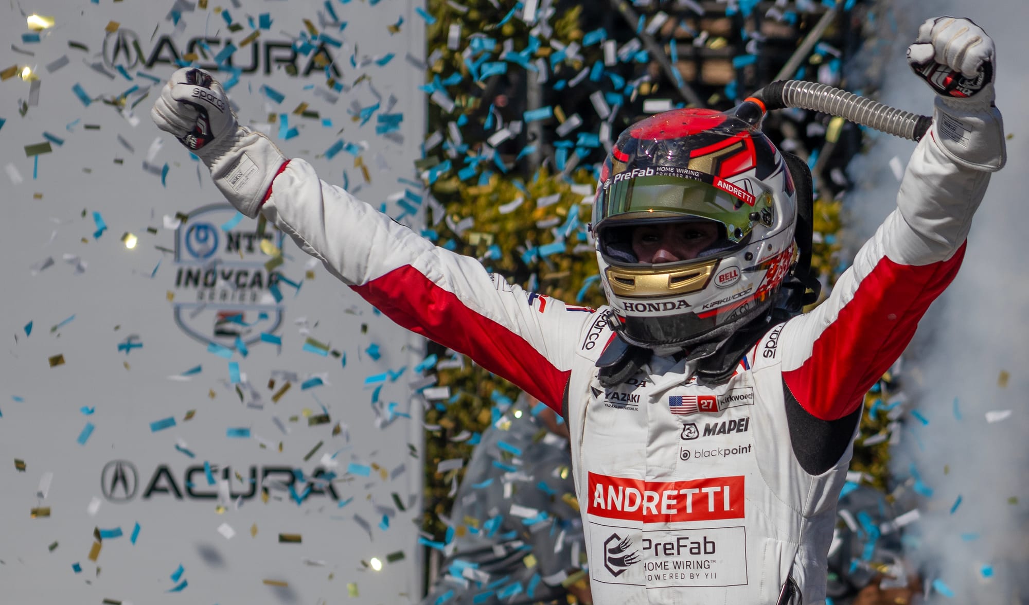 A man in a racing helmet holds up his arms as confetti rains down.