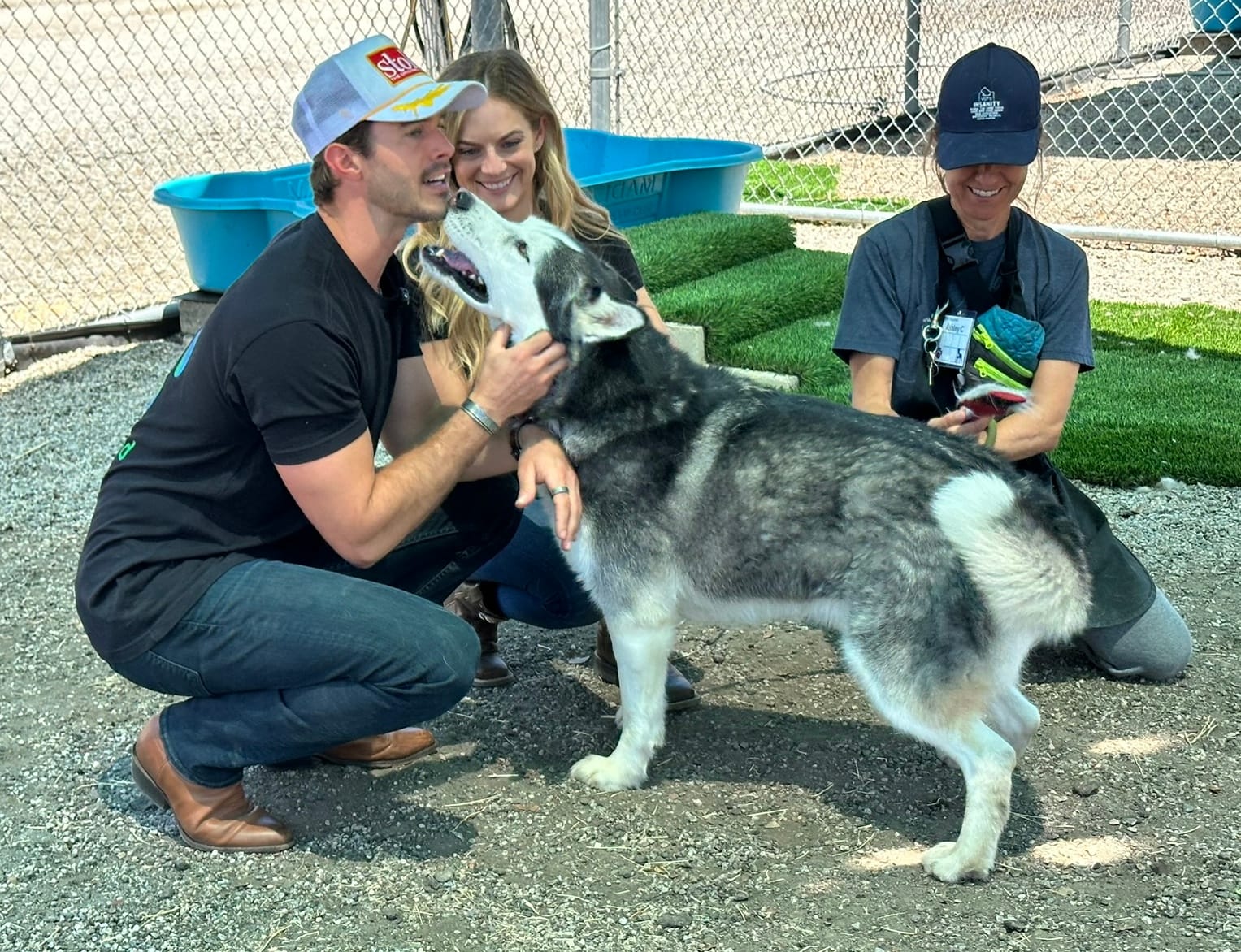 A white and gray dog licks a man's face while two women kneeling nearby smile.