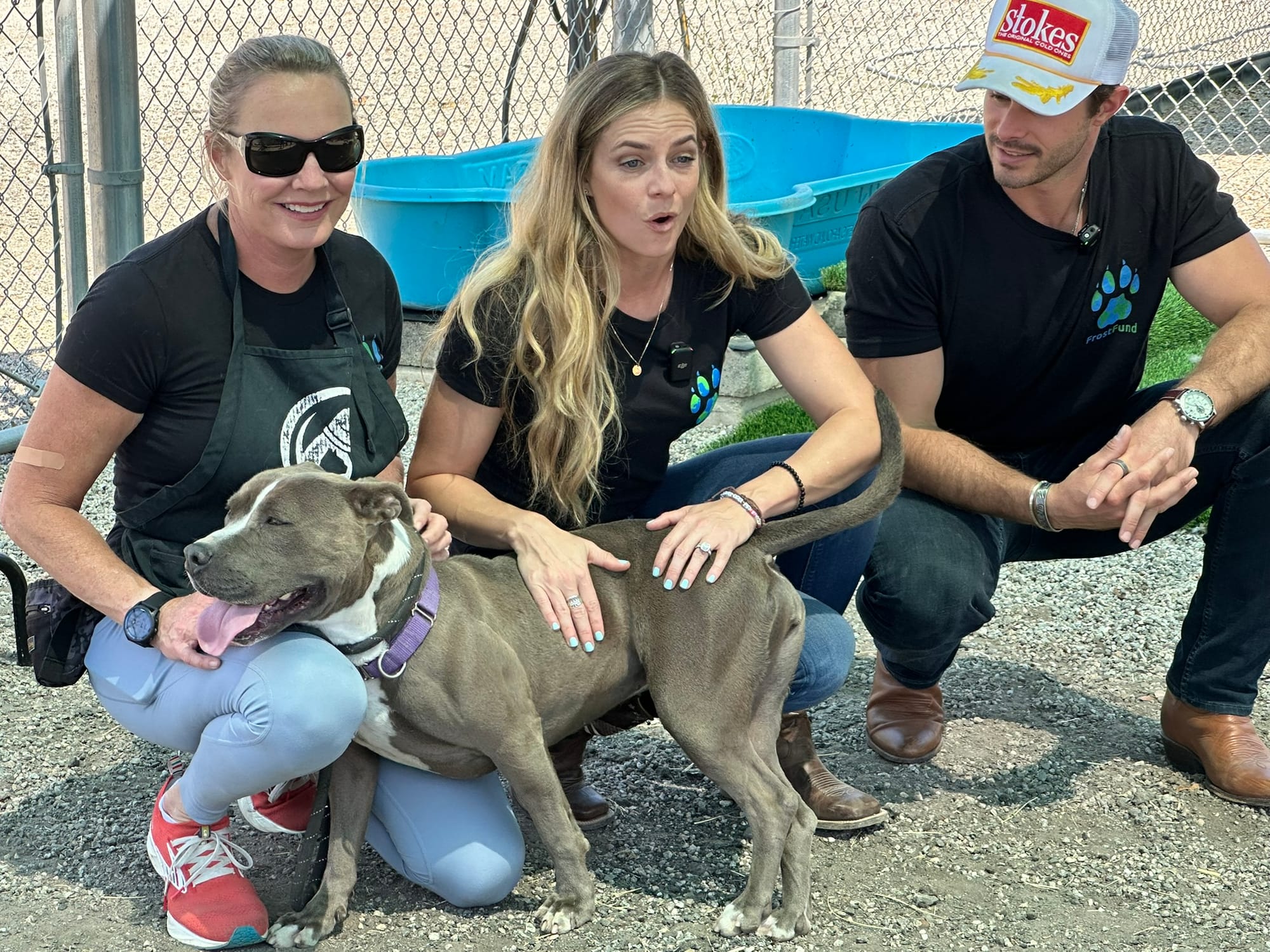 Two women and a man kneel on the ground with a sweet dog.