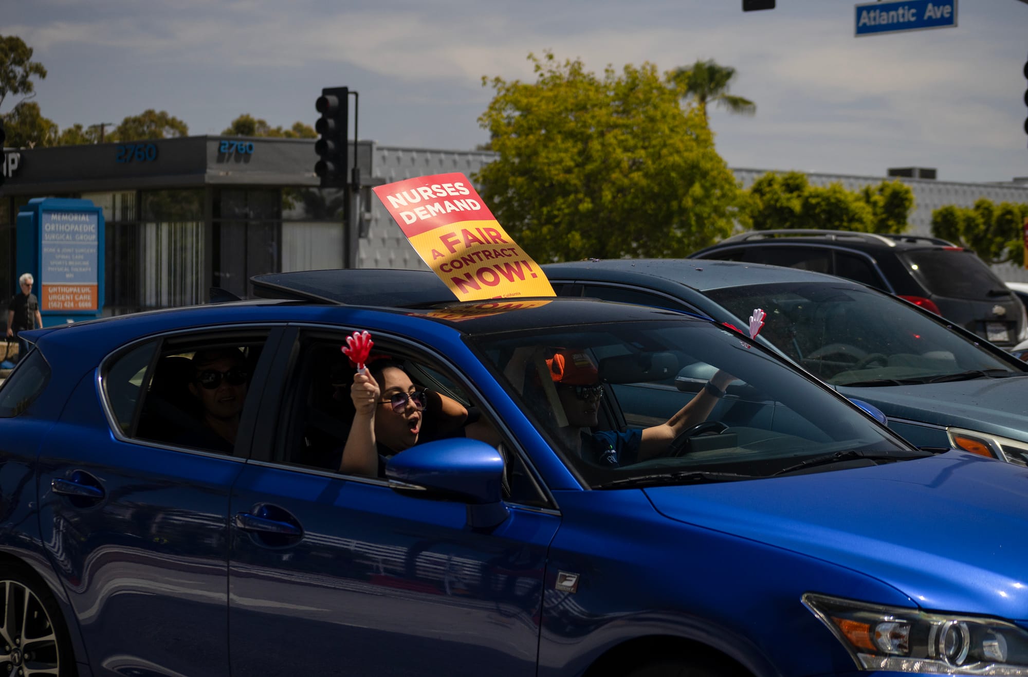 A person in a blue car holds a sign saying "nurses demand a fair contract now!" through the sun roof.