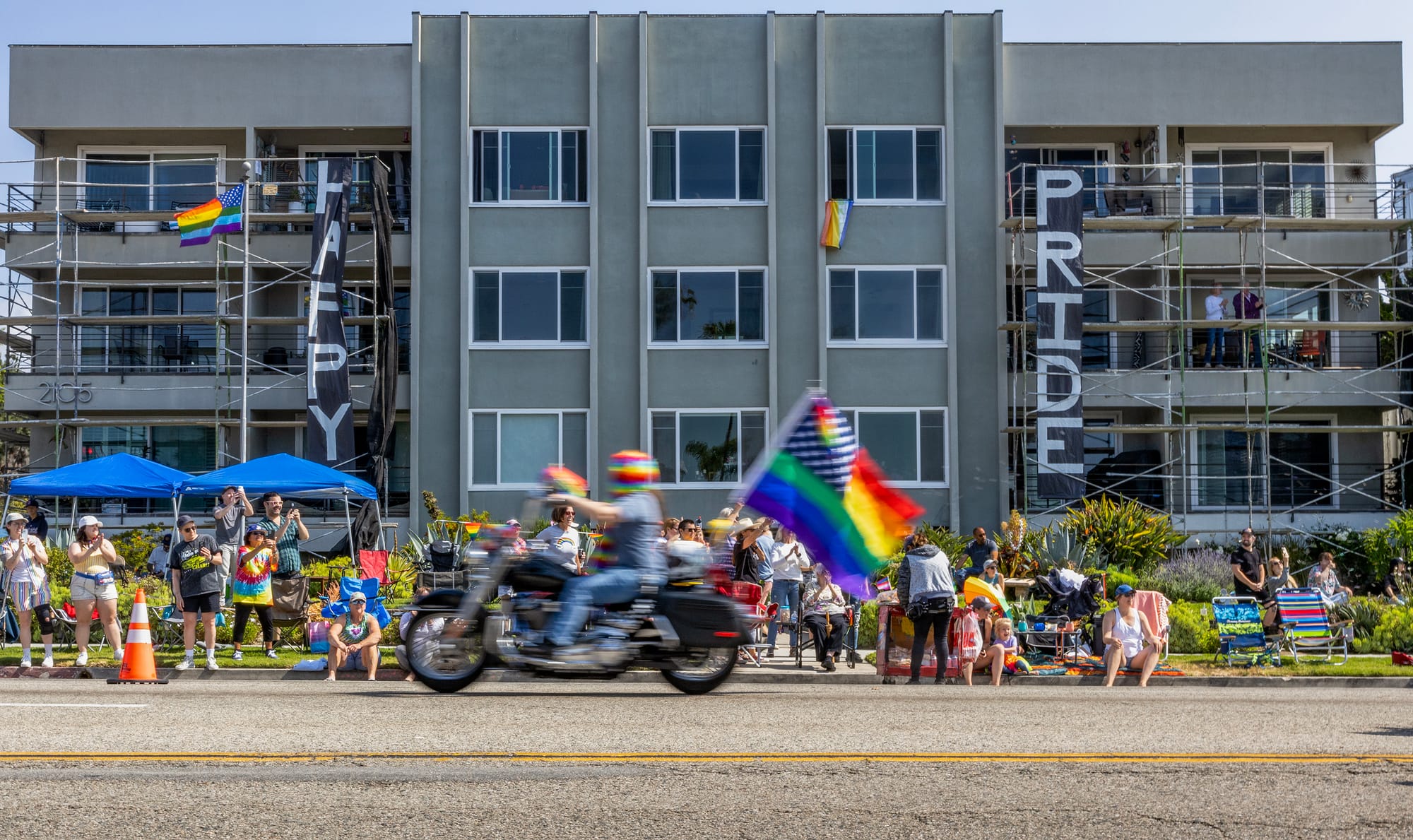 Blurry image of a motorcycle with a rainbow-colored US flag riding down the street in front of a gray building.