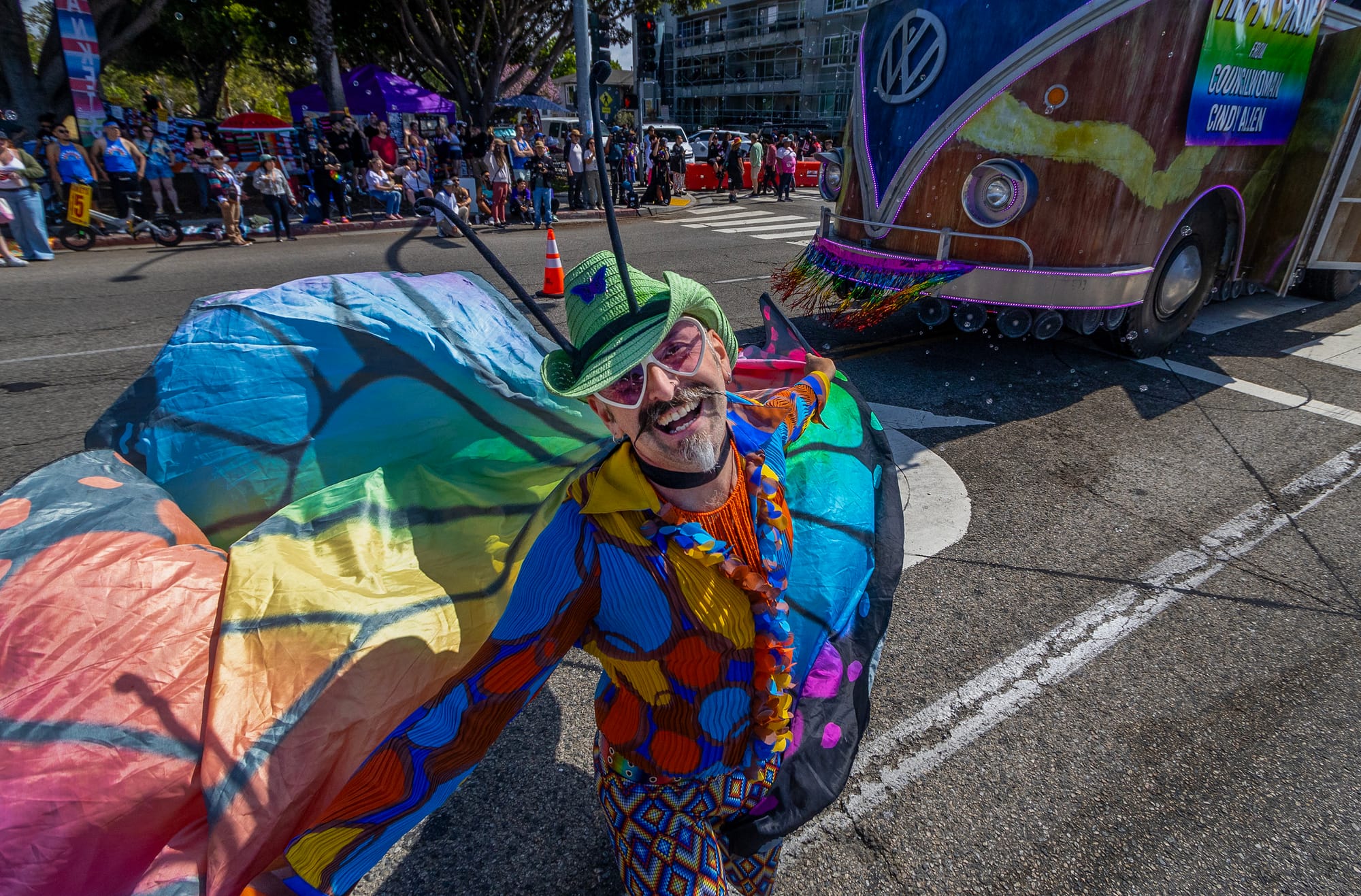 A man wearing colorful clothes and a rainbow-colored cape smiles in the middle of a street.