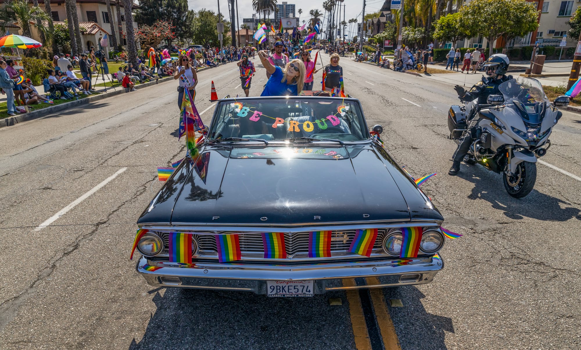 A woman sits in a convertible that has the words "be proud" stretched across the windshield taking a selfie.