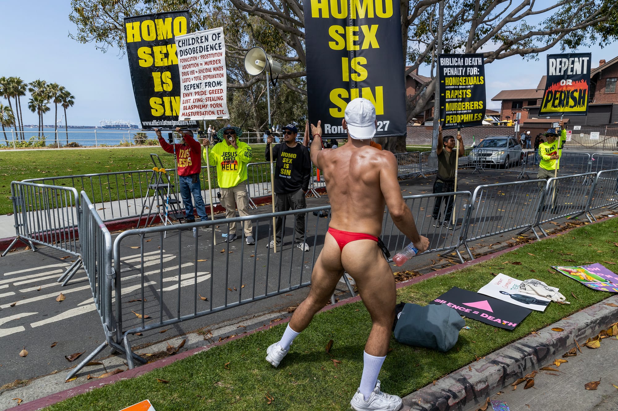 A man wearing only white shoes and a red speedo swimsuit stands in front of people holding signs saying "homo sex is sin."
