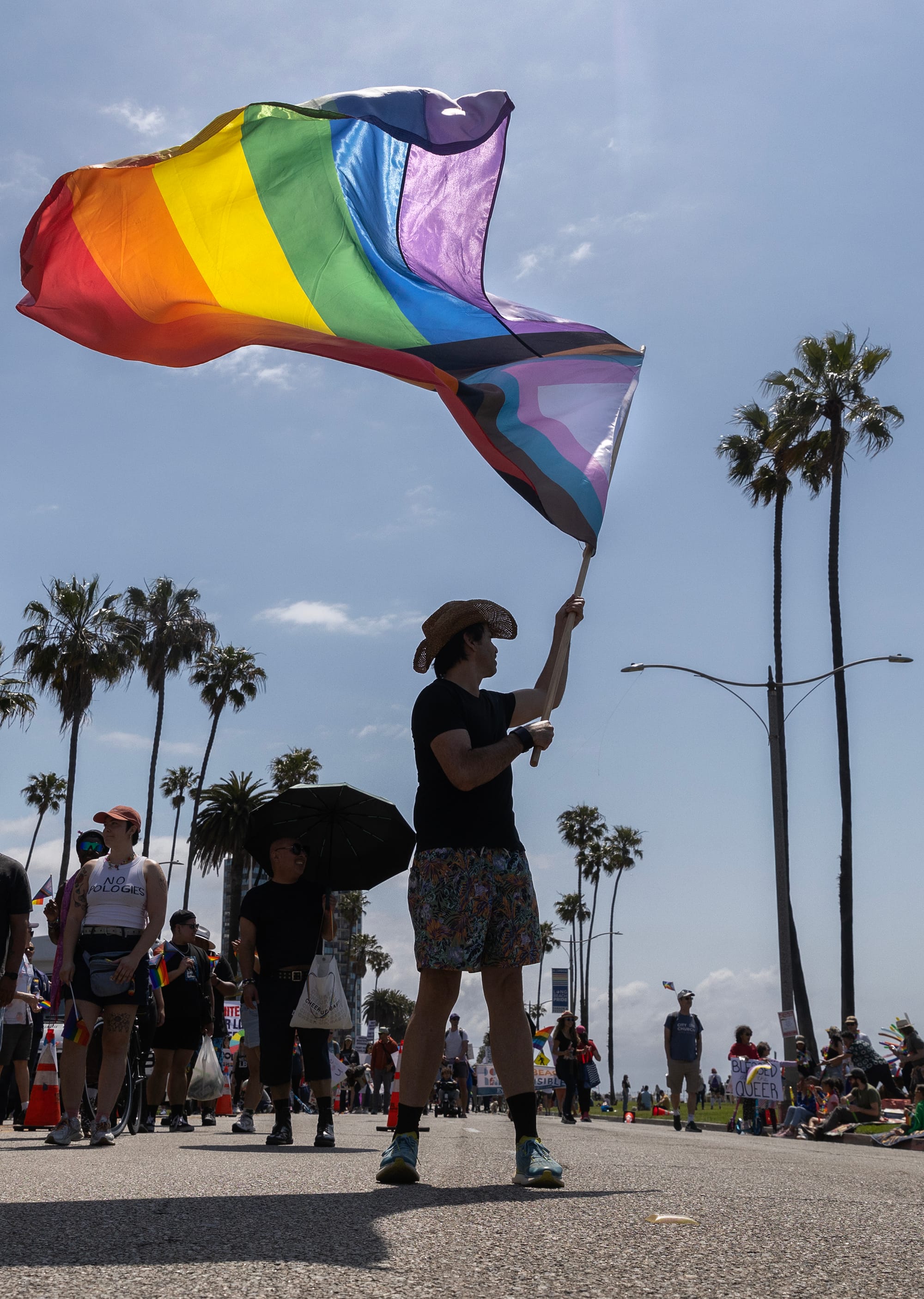 A man in a cowboy hat stands in the street waving a rainbow flag.